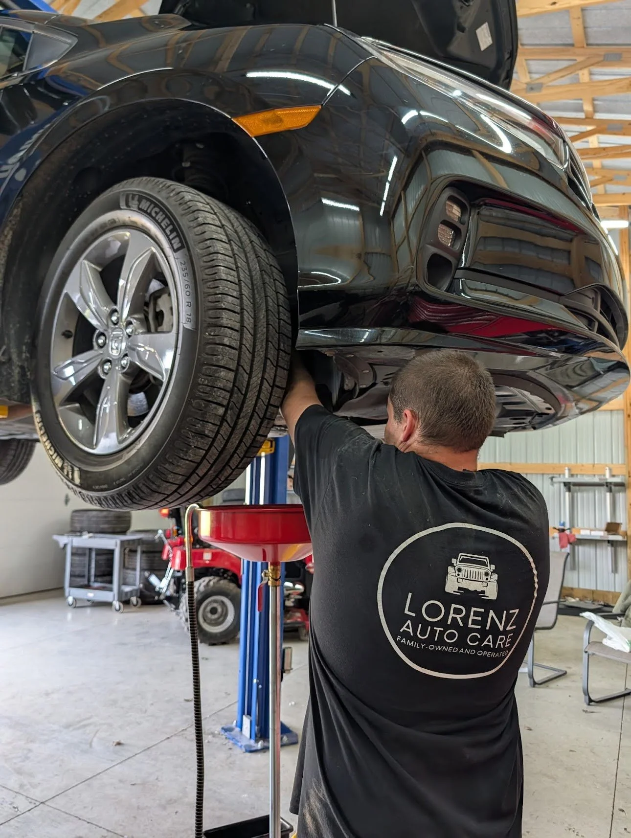 A mechanic working on a black car elevated on a hydraulic lift inside an auto repair shop. The mechanic is wearing a black t-shirt with 'Lorenz Auto Care' on the back. 