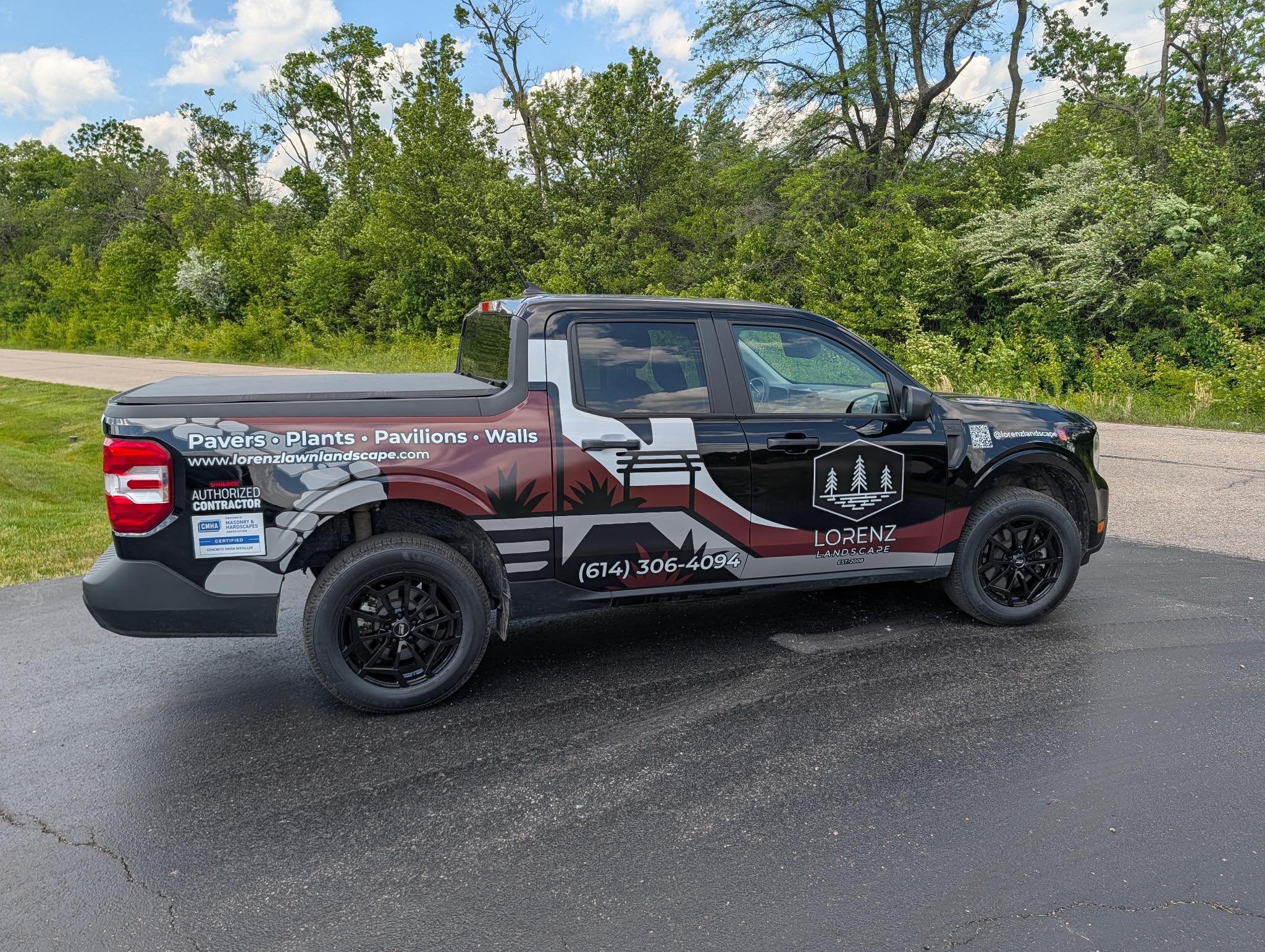 A black and gray pickup truck with advertisements for a landscaping business on its side, parked on a paved surface with green trees and a partly cloudy sky in the background.