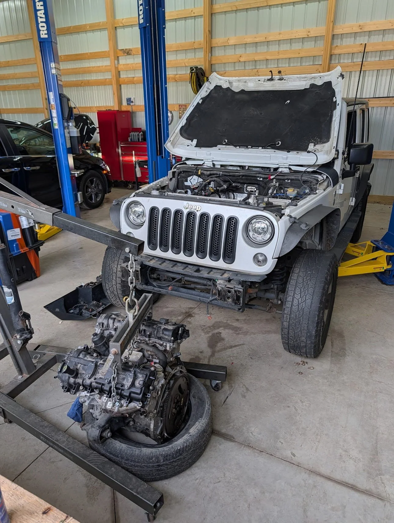 A white Jeep with its hood open in a garage, with an engine and transmission removed and placed on a frame