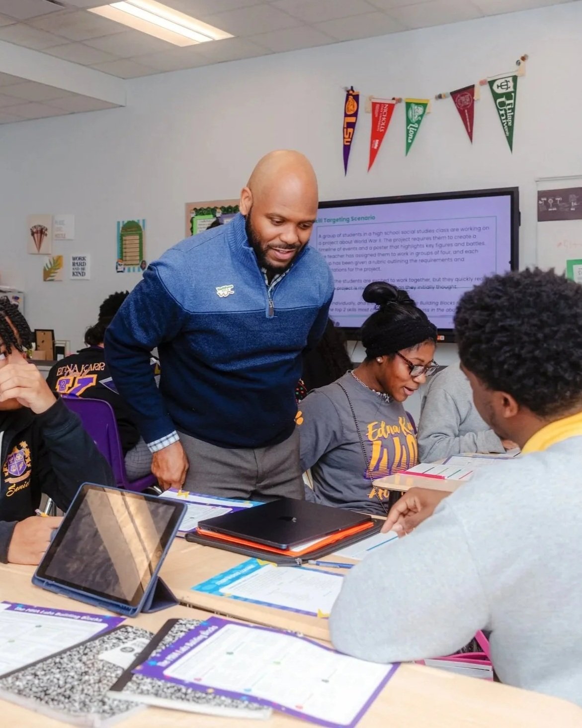 Smiling teacher stands among a group of students sitting at desks looking at their work