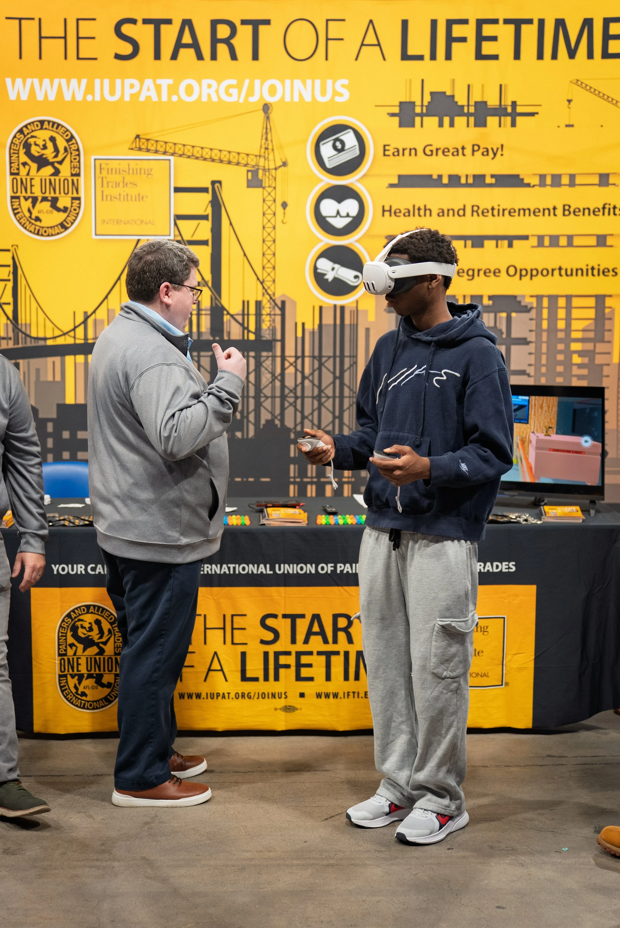 A man and a student stand in front of a yellow backdrop depicting a construction site. The backdrop reads "The Start of a Lifetime." The student is wearing a VR headset.
