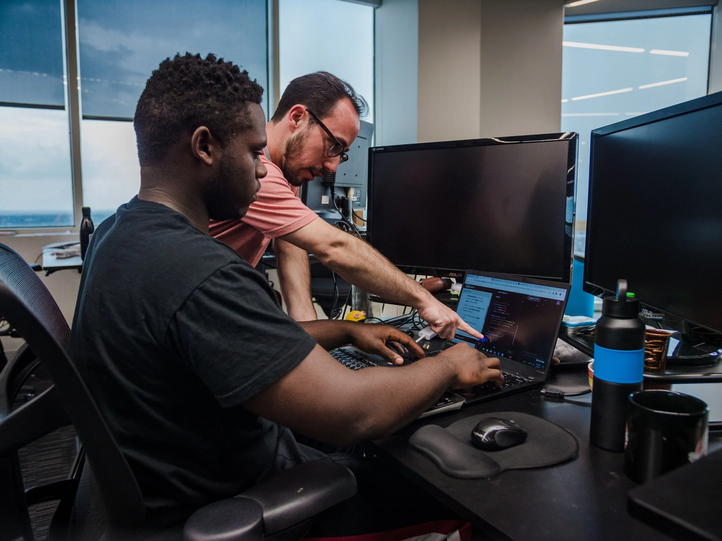 A man in a pink shirt is standing behind a student sitting at a computer; he is pointing at something on a computer screen that is front of them both.
