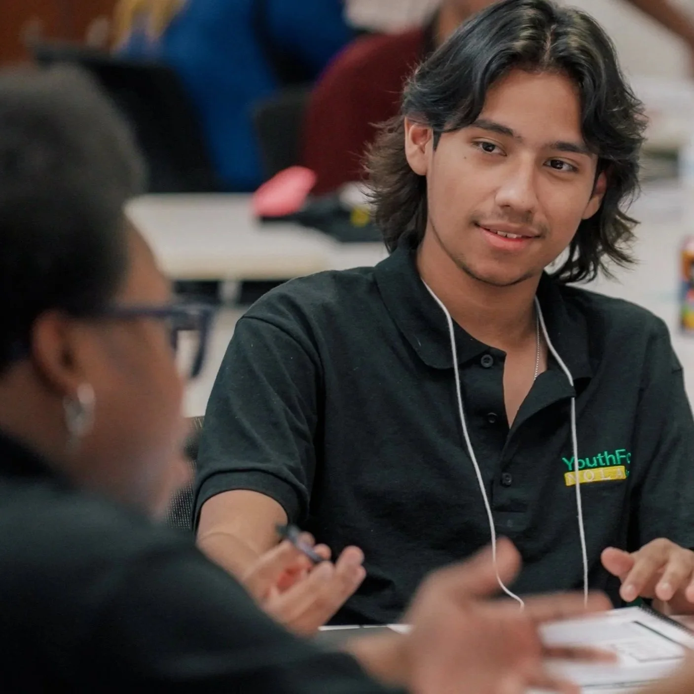 Hispanic-appearing boy sits at a table and listens to the woman in front of him. He is wearing a black Youth Force NOLA shirt.