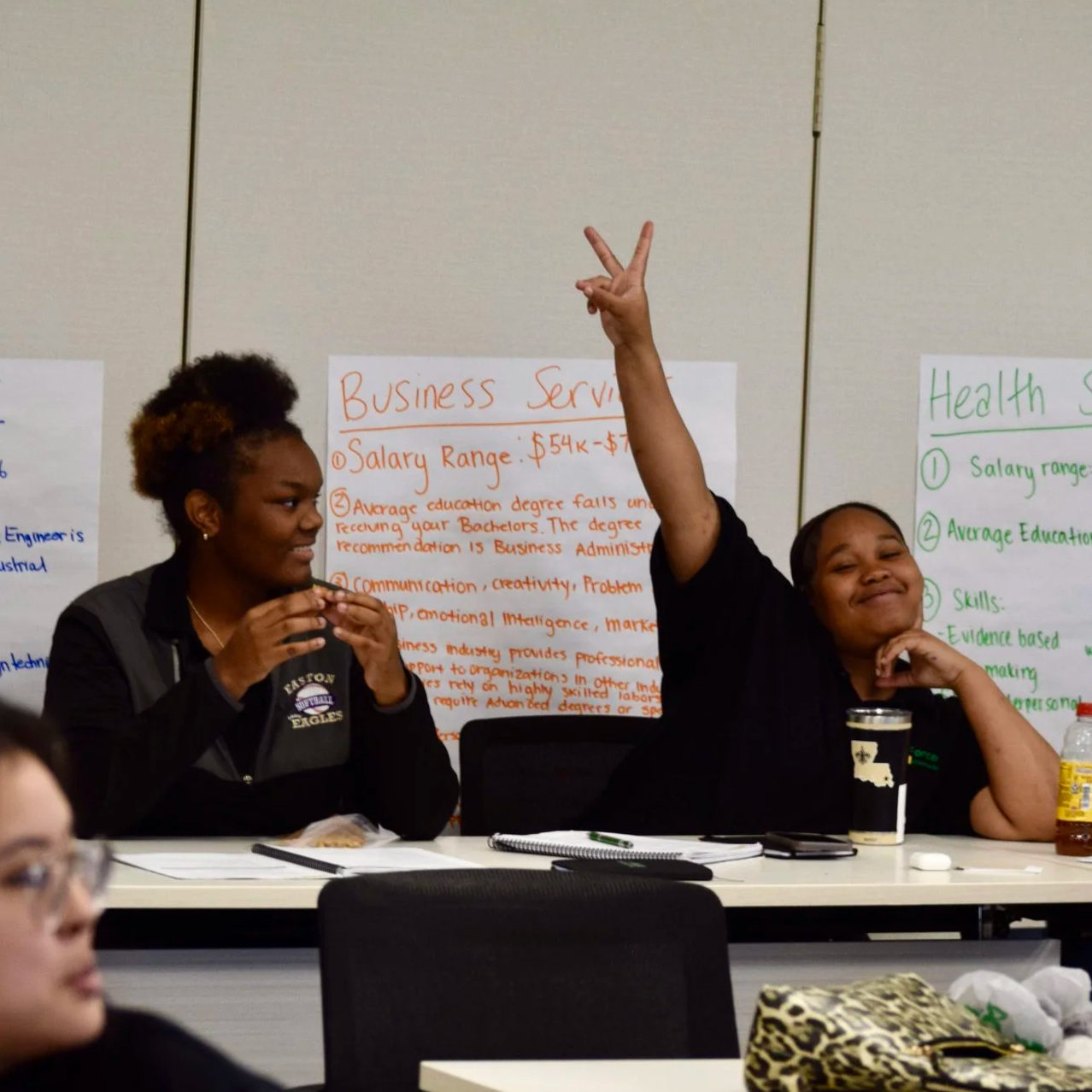 Two black female students sit in a classroom smiling. One has her hand up in a peace sign.
