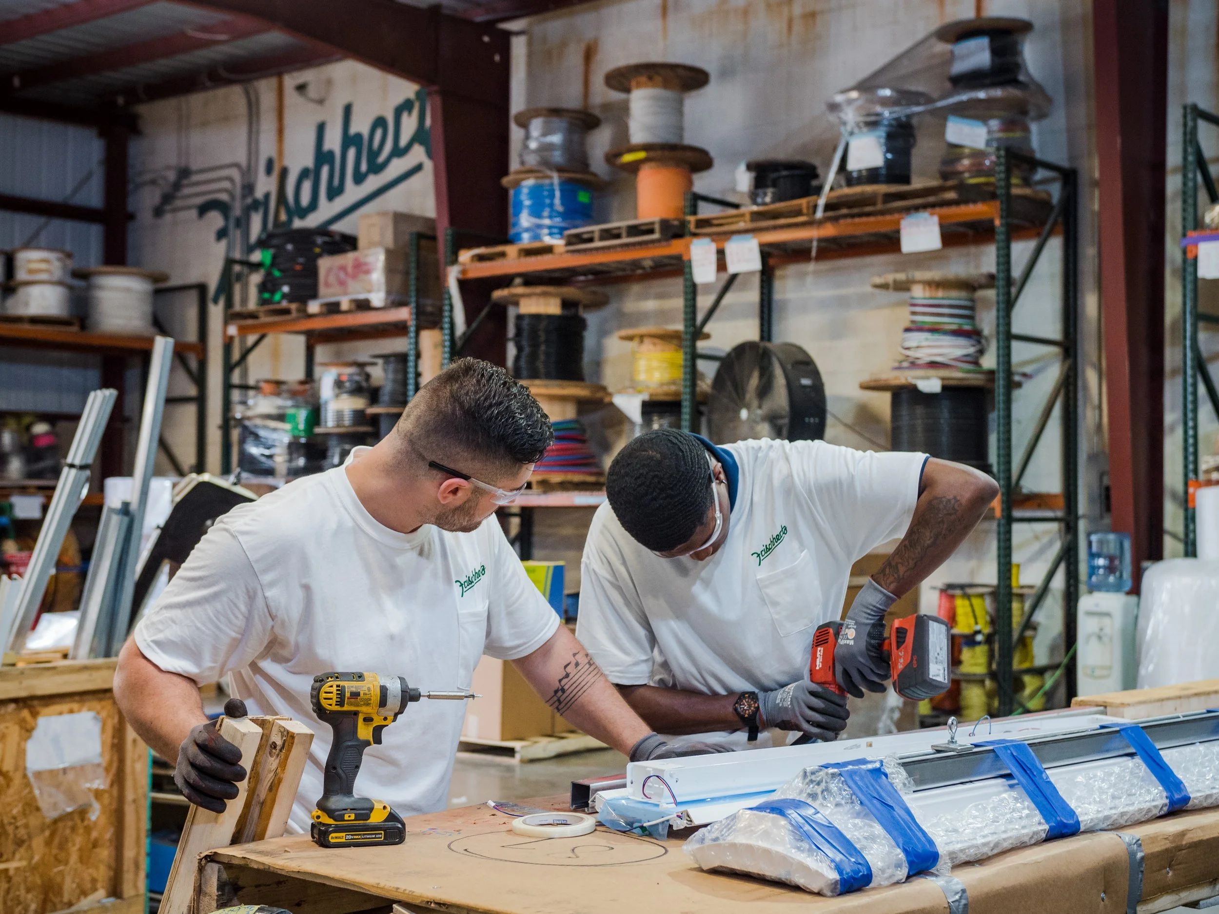 Two men in white shirts stand behind a carpentry bench; one is using a drill.