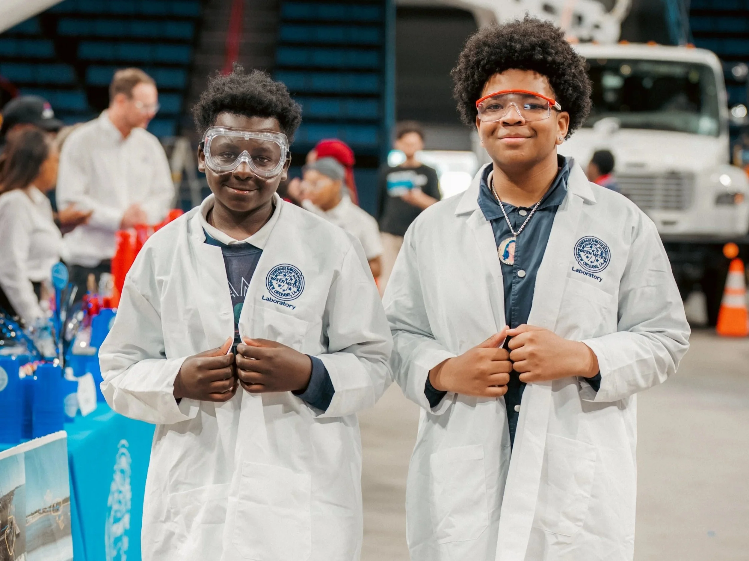 Two black boys stand smiling, side-by-side in white lab coats with a blue logo and safety glasses