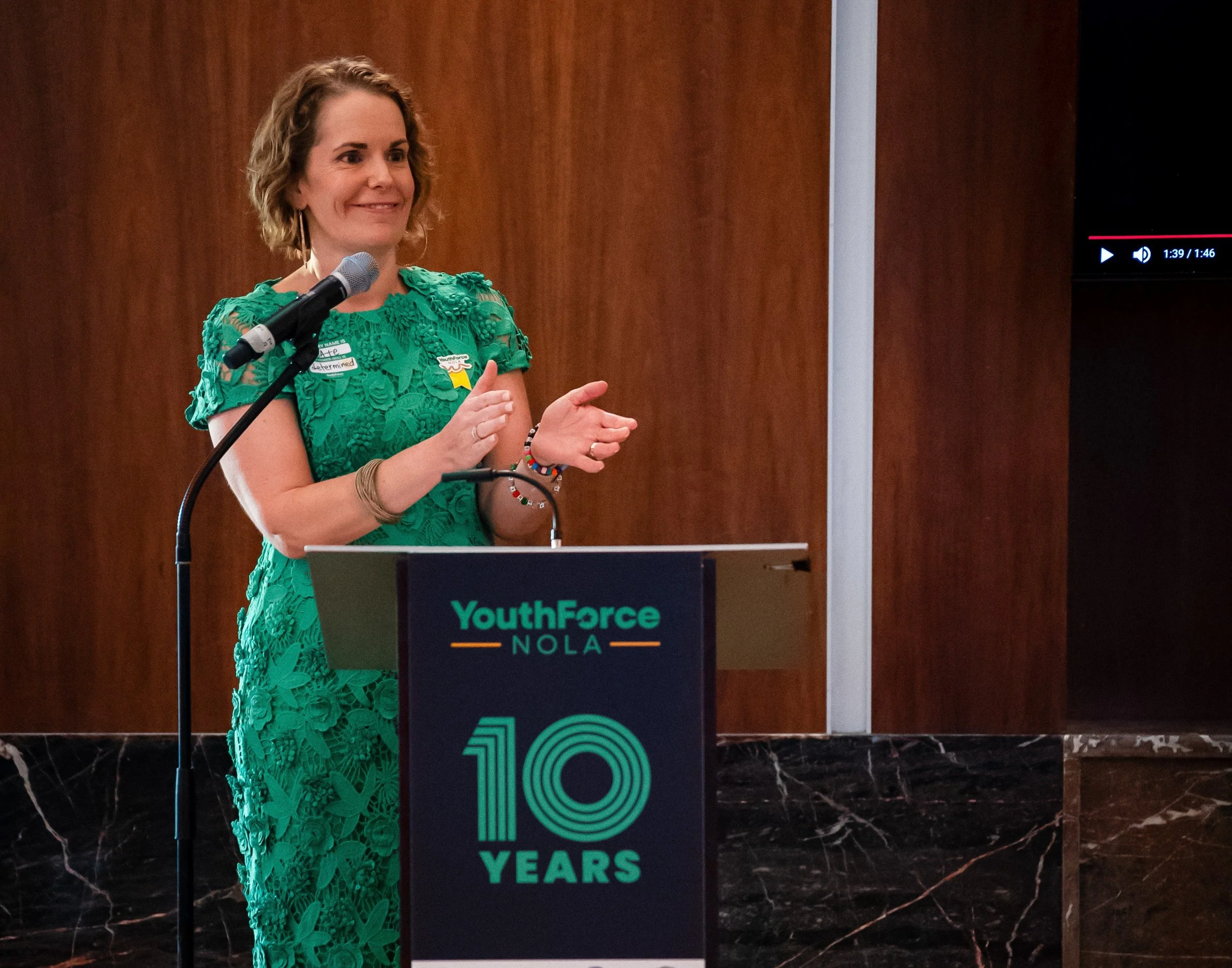 YouthForce NOLA President and CEO Cate Swinburn stands behind a podium; she's wearing a green dress and clapping in celebration of YouthForce NOLA's 10th Anniversary.