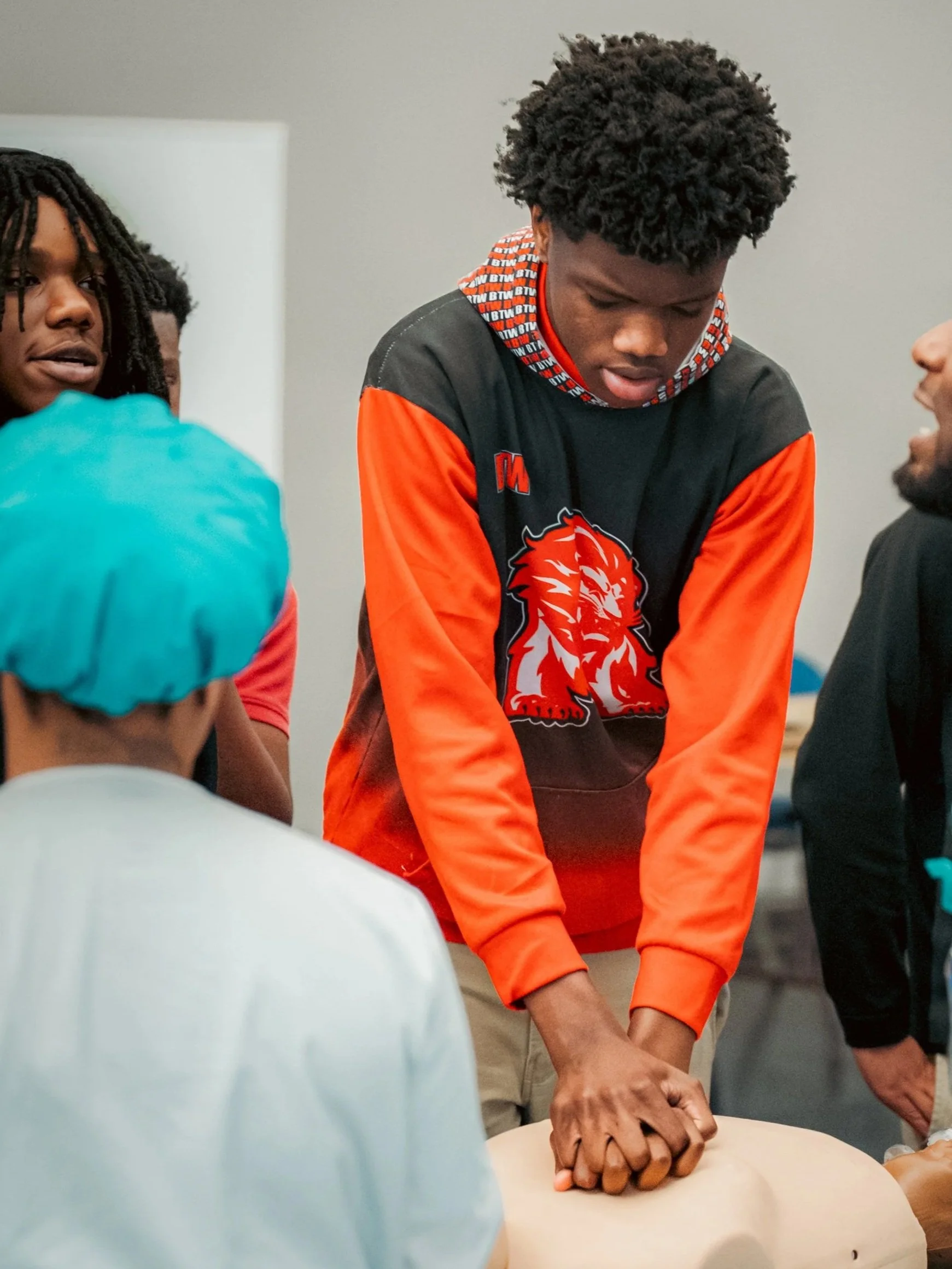 A black boy in a black and orange hoodie practices CPR on a dummy.