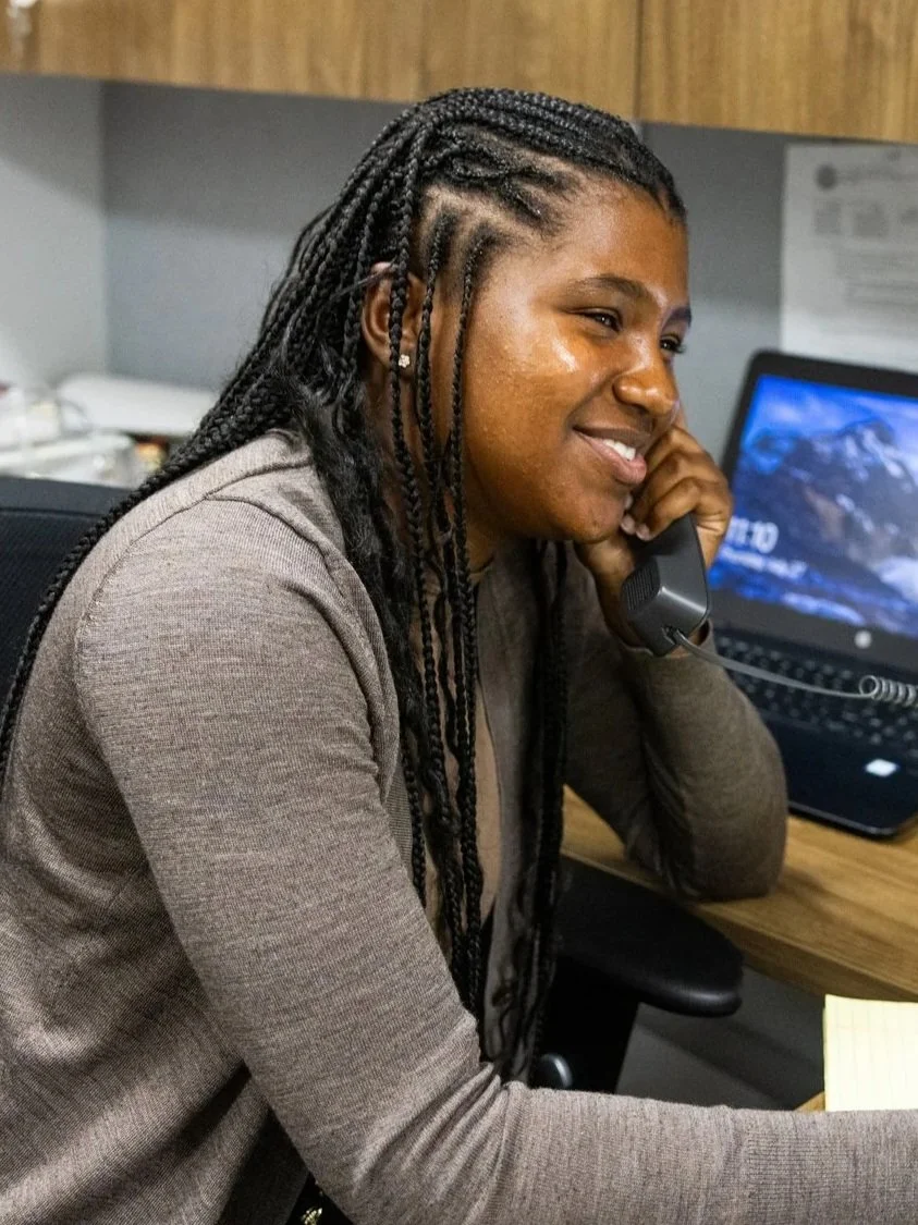 A black girl sits behind a desk. She is smiling and holding an office phone to her ear.