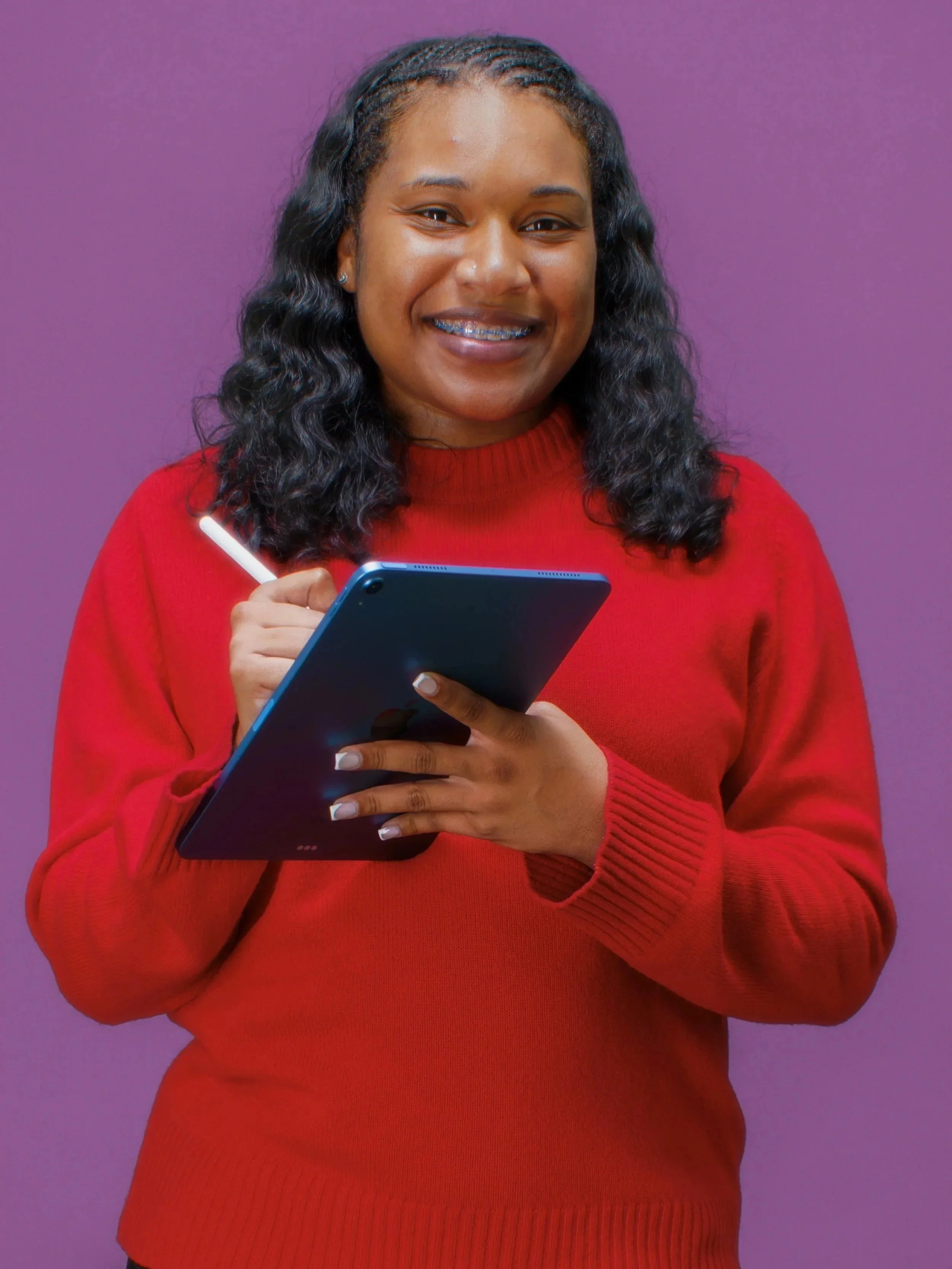 Tatyana Stevenson stands smiling in front of a purple background holding an ipad. She is wearing a red sweater shirt.