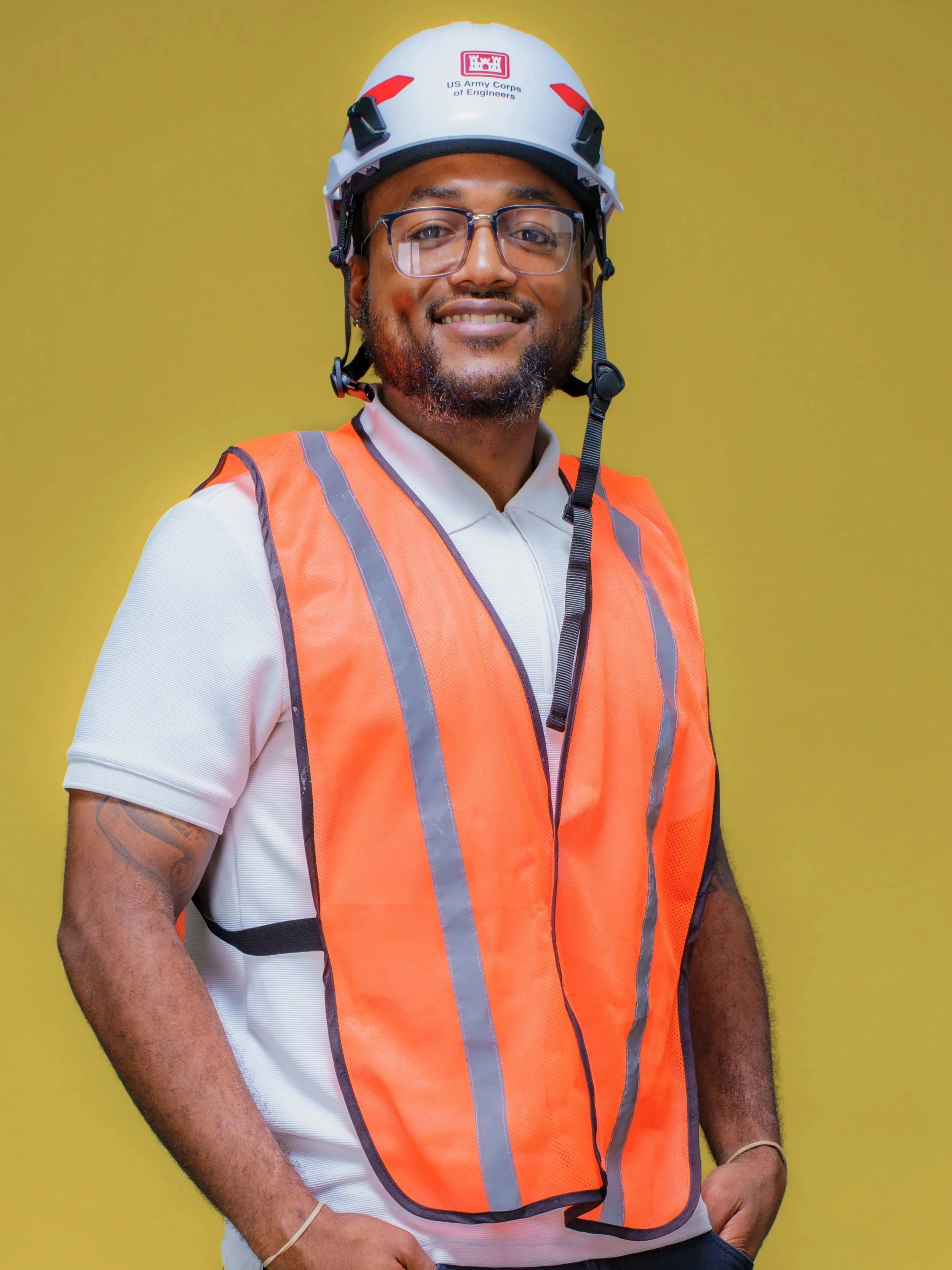 Nathanial Kenner is wearing a white shirt under an orange safety vest and white helmet with the US Army Corps of Engineering logo on it. He is standing smiling in front of a yellow background.