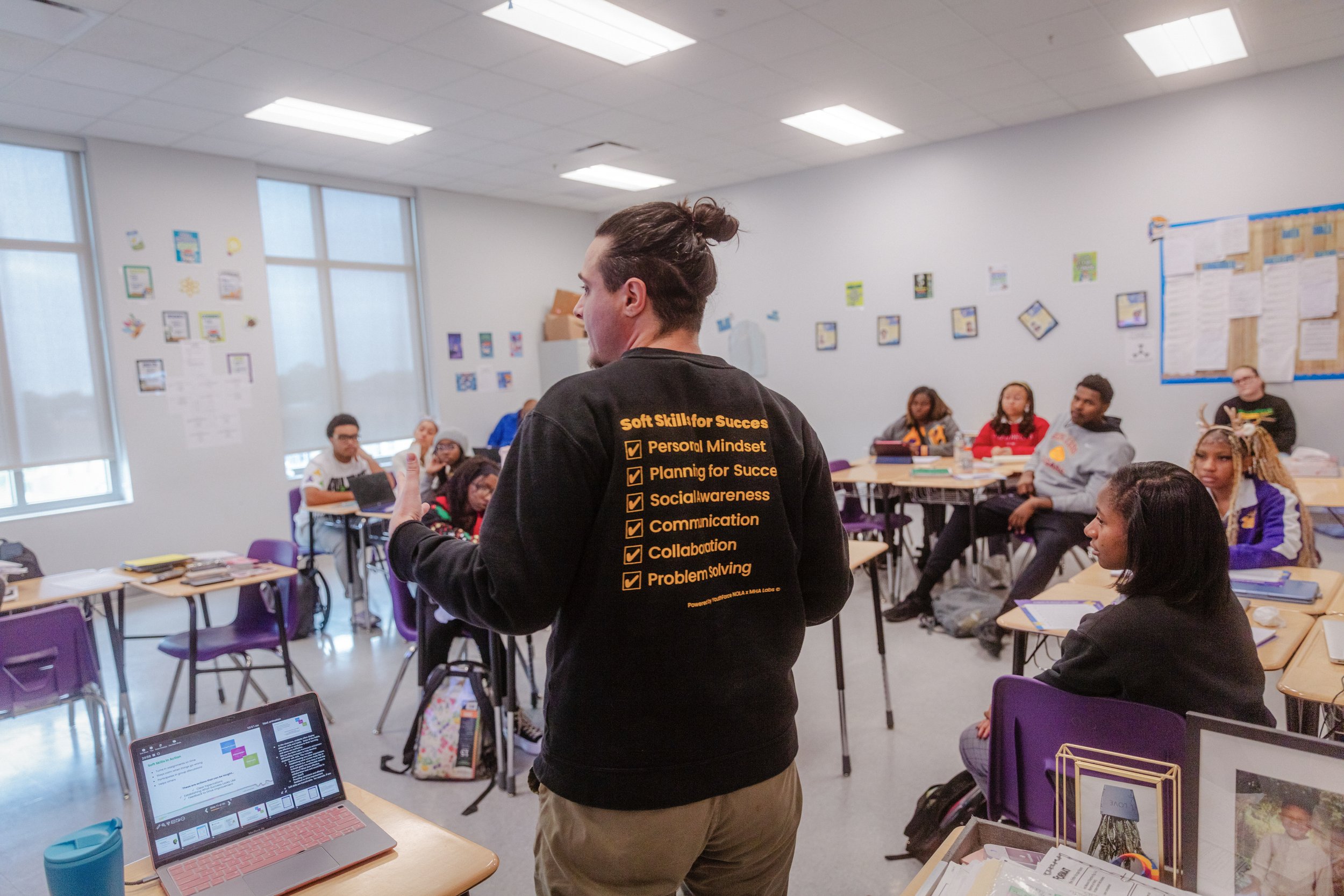 A man stands in front of a classroom with his back to the camera. He is wearing a black shirt that has a checklist of soft skills for success written in yellow.