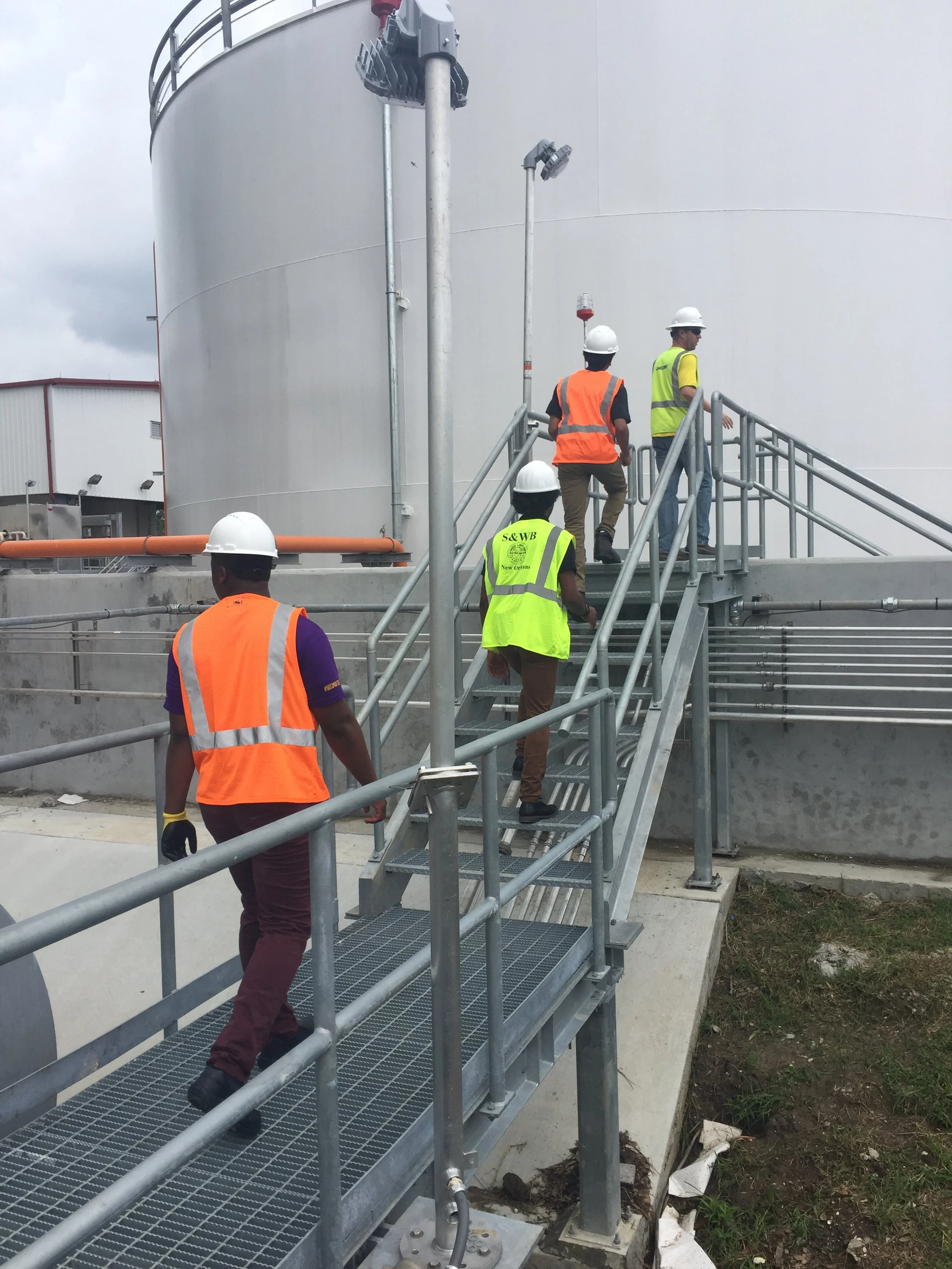 Four people in yellow and orange safety vests and white helmets are walking away from the camera along a metal walkway.