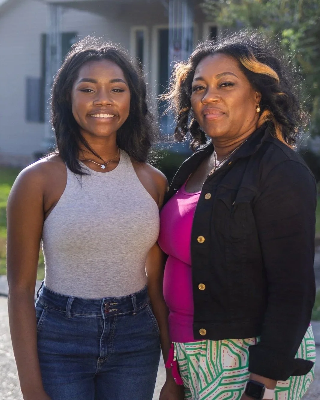 Image of a black mom and daughter smiling at the camera; a house is in the background