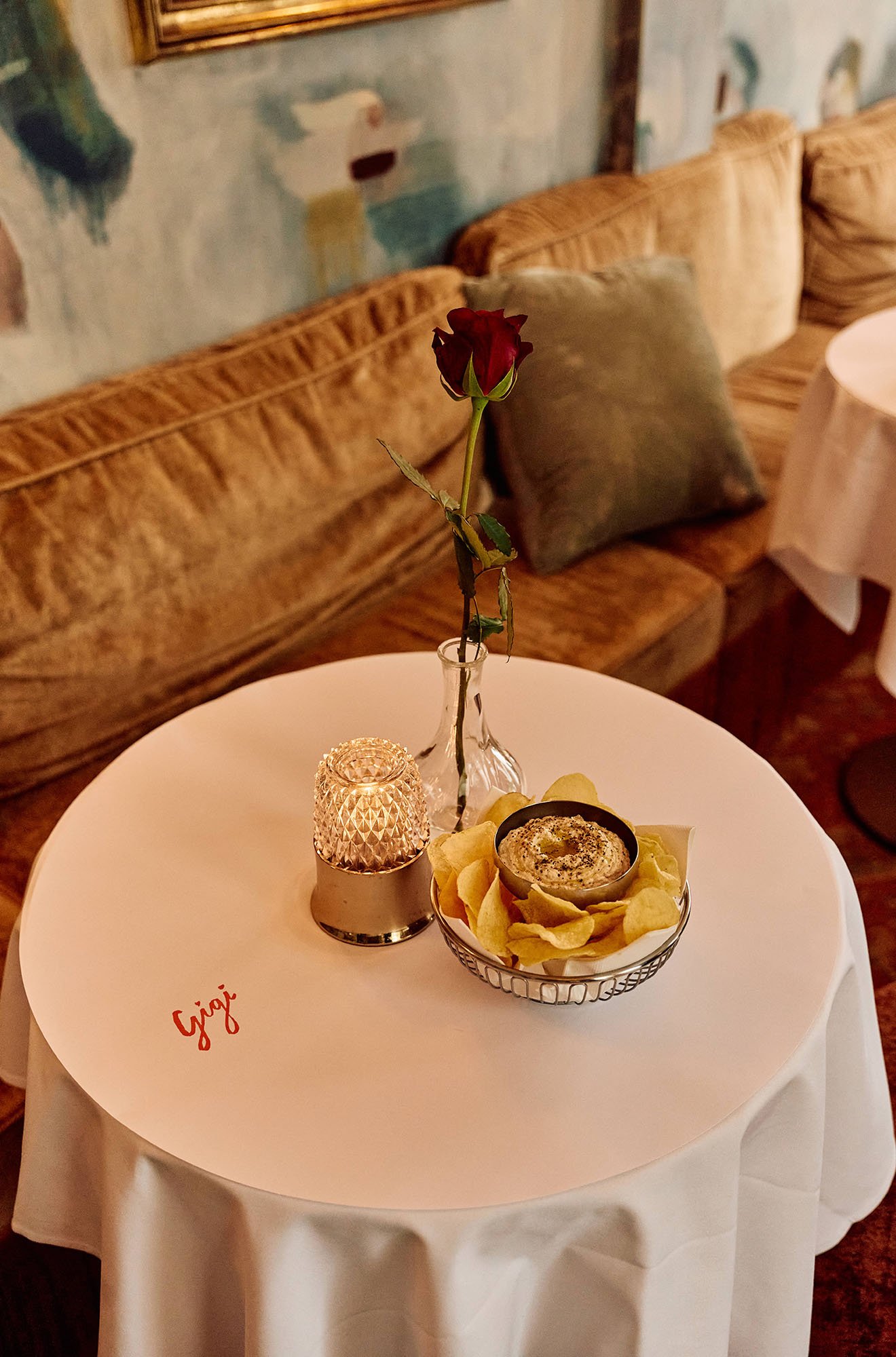 A small round table covered with a white tablecloth. On the table, there's a single red rose in a clear glass vase, a small decorative candle holder, and a bowl of chips with a creamy dip. The table has the word 'Gigi' written in red cursive in the l