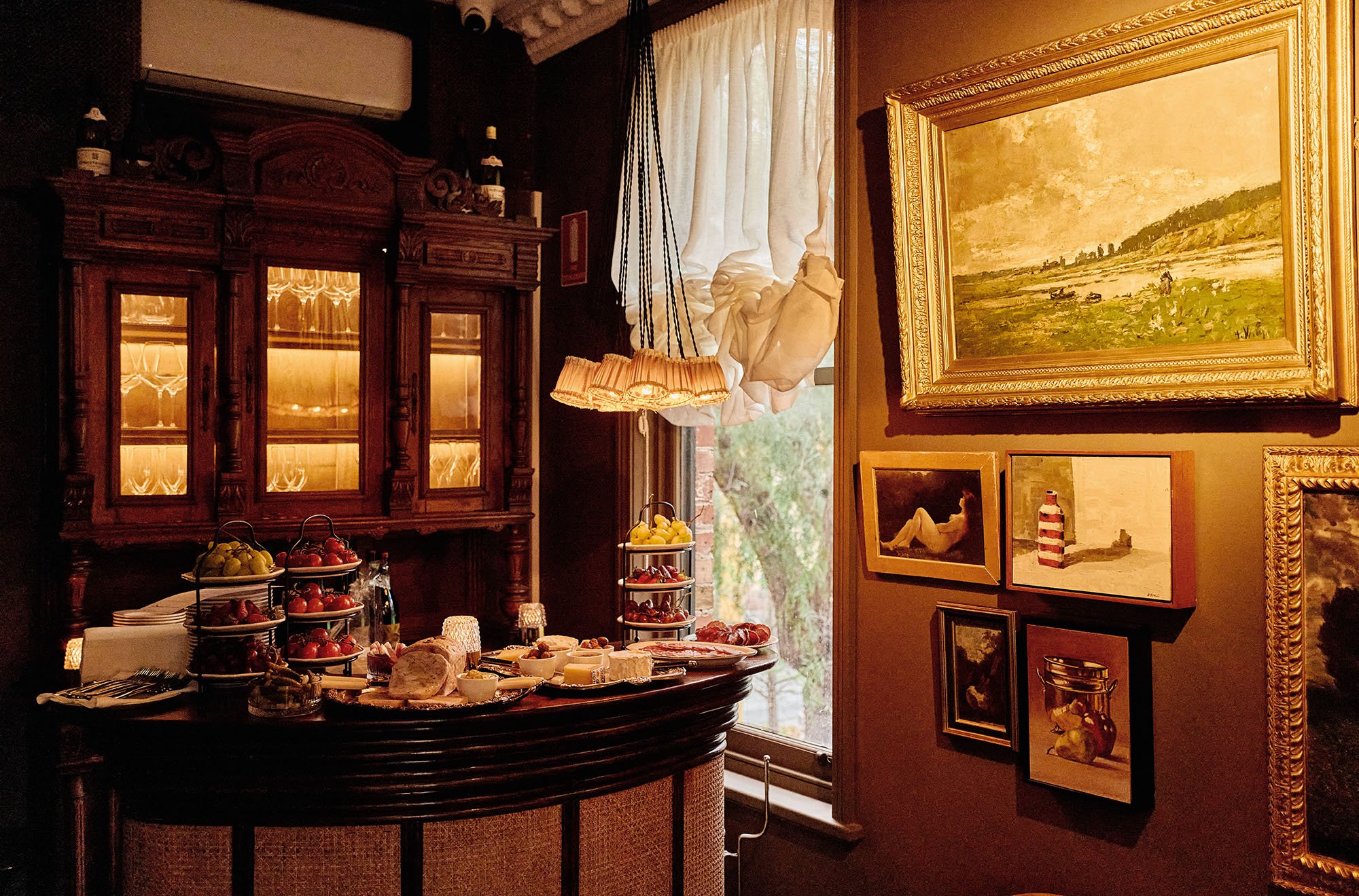 A cozy room with a display of food and art. There are several pieces of framed artwork on the wall, a wooden cabinet with glass doors, a window with white curtains, a hanging lamp with a fabric shade, and a table filled with bowls of fruit, cheese, a
