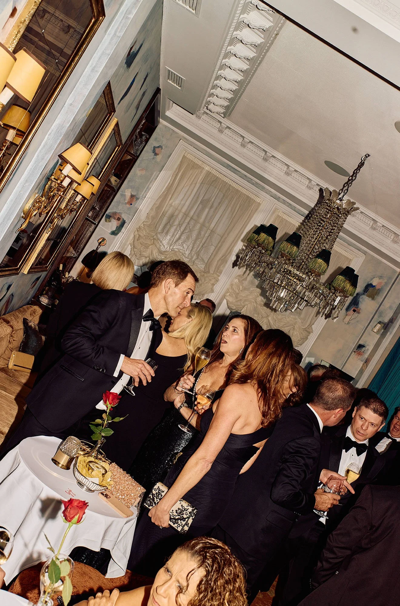 An elegant party or gathering in a decorated room with chandelier lighting, intricate crown molding, and artwork on the walls. People dressed in formal attire, including tuxedos and evening gowns, are holding drinks and socializing. A table decorated
