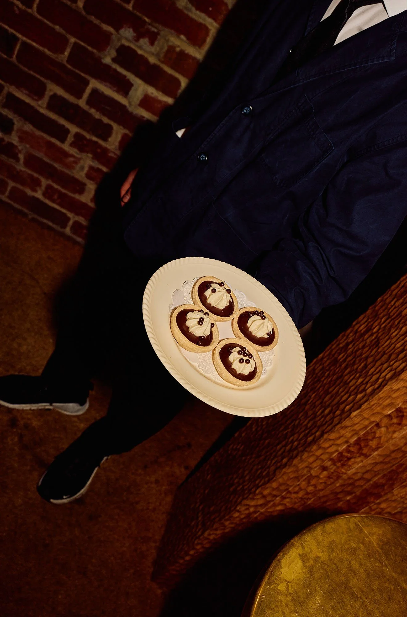 Person holding a white plate with five decorated cookies. The cookies are shaped like Santa Claus with white icing, chocolate, and candy decorations. The person is wearing a dark jacket, black pants, and black sneakers with white soles, sitting in a 
