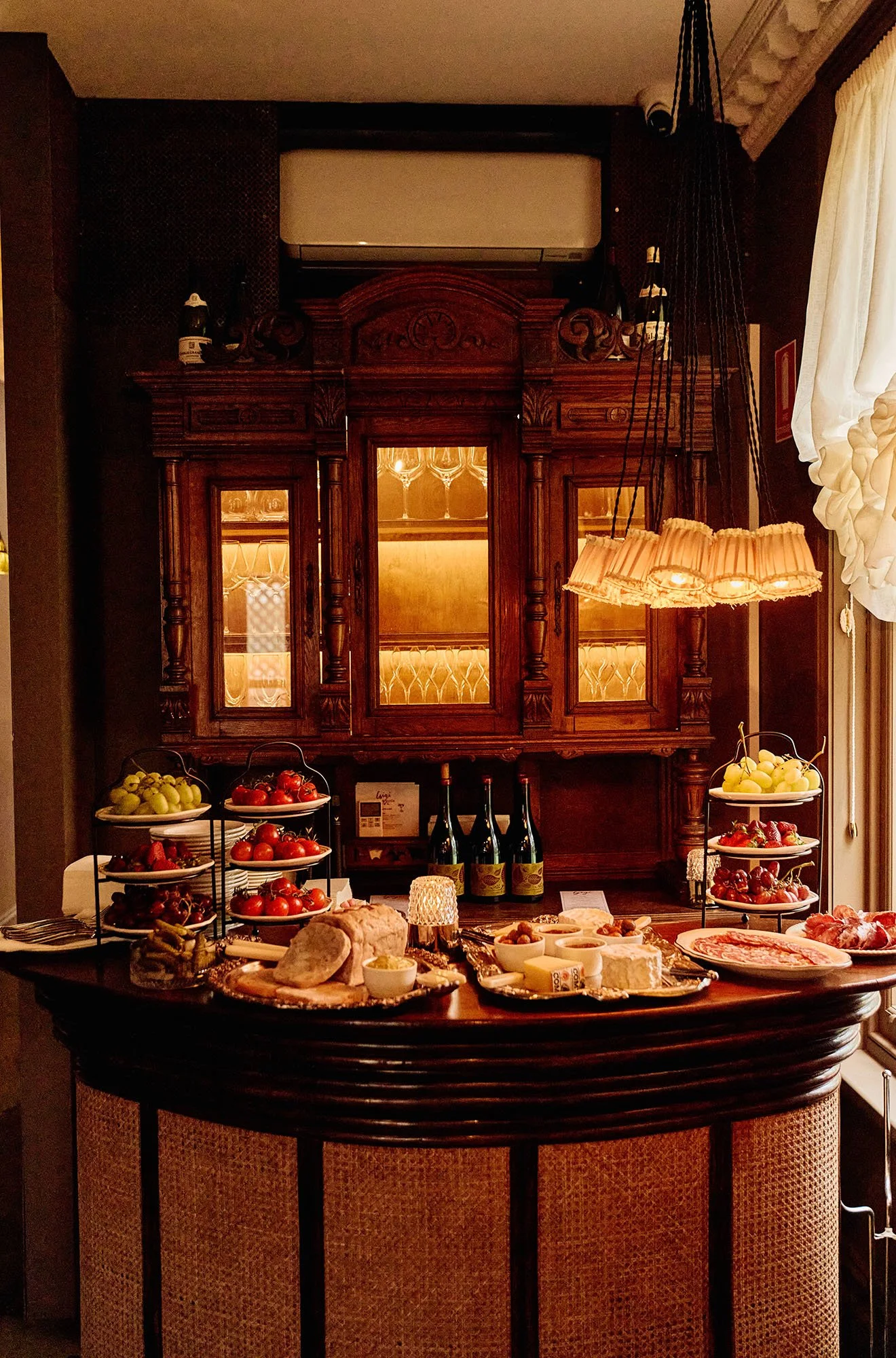 A dining table set with various cheeses, meats, bread, and fruits, with tiered fruit trays and wine bottles in front of an ornate wooden cabinet and a warm lighting interior.