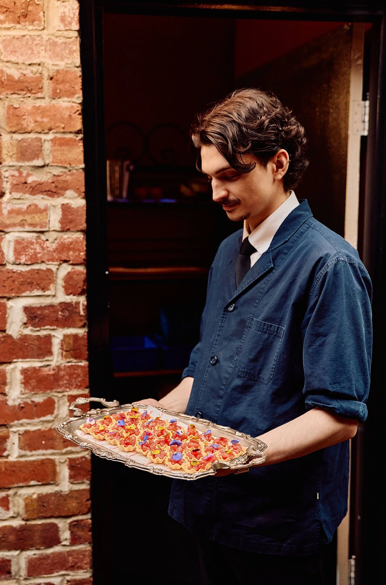 A man in a blue jacket and white shirt holding a tray of colorful decorated cookies or dessert items, standing beside a brick wall in an indoor setting.