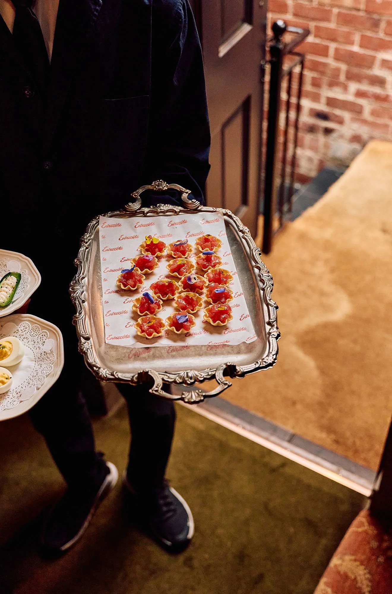 A person wearing dark clothing and black sneakers is holding a silver tray with mini tartlets topped with red fruit, possibly strawberries and blueberries, and garnished with edible flowers. The tray has ornate edges and white paper liner with red te