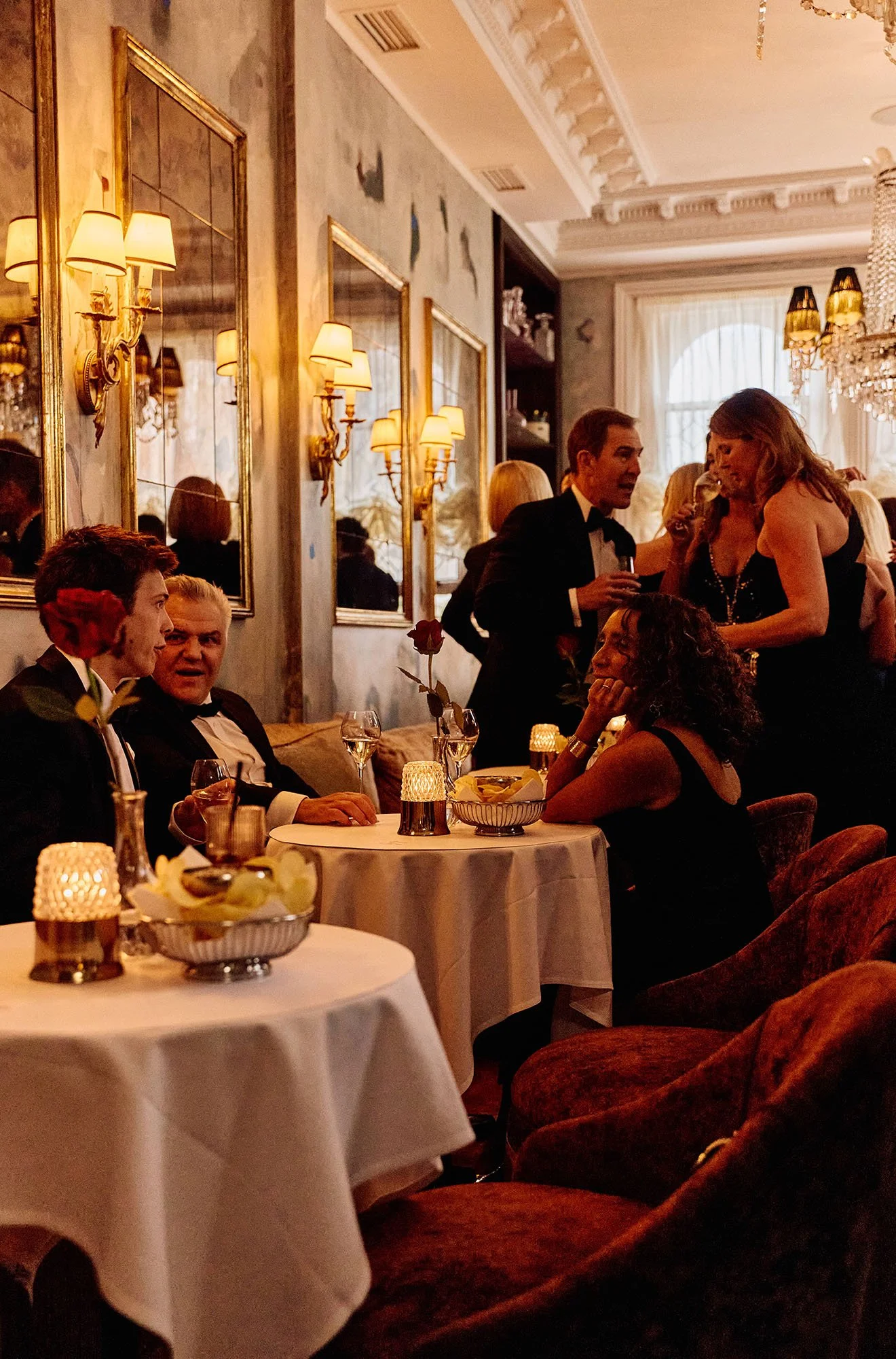 A group of people dressed in formal attire enjoying a dinner and conversation in an elegant, warmly-lit restaurant with ornate decor and wall sconces.