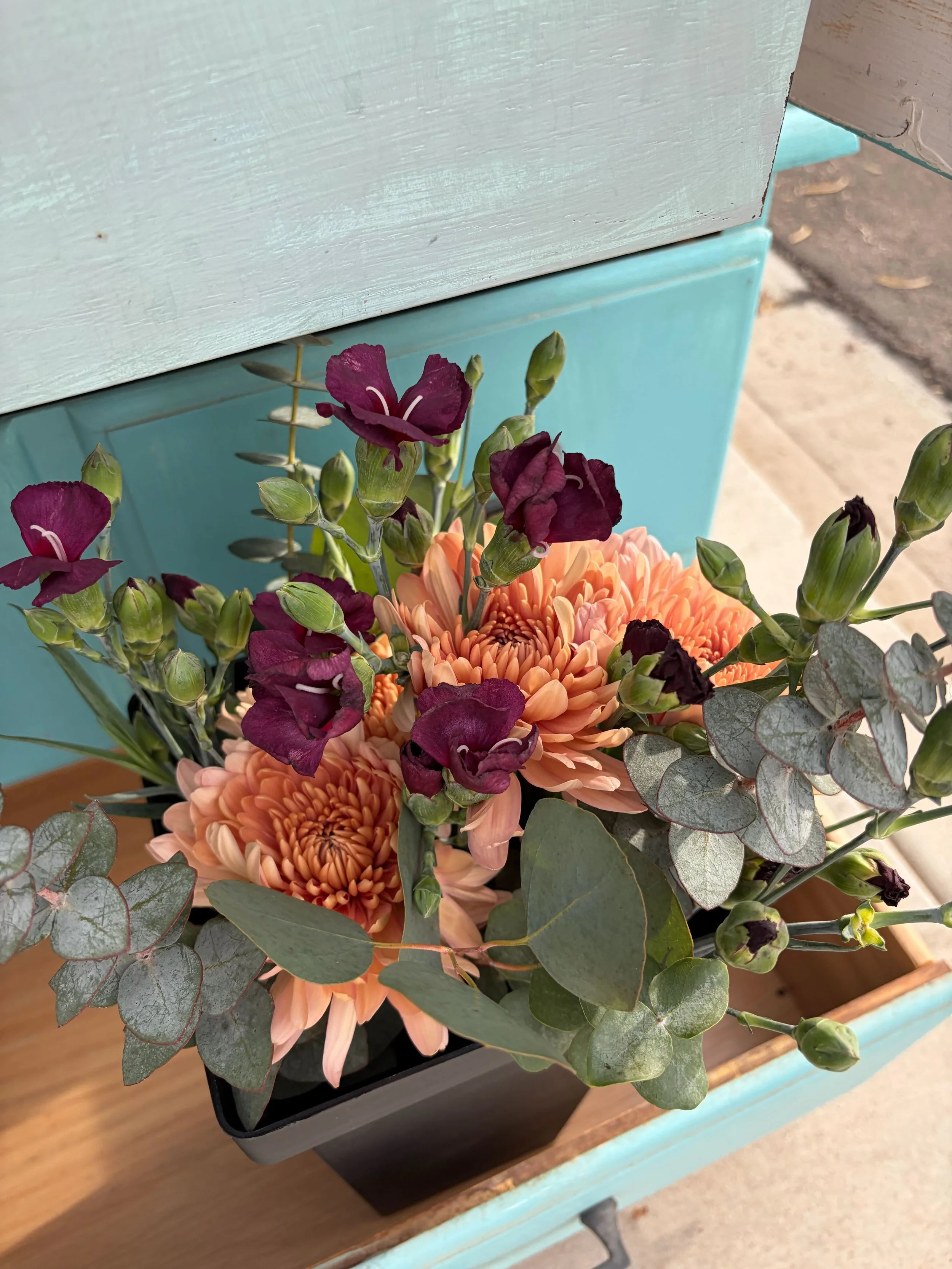 A bouquet of peach-colored flowers with purple blooms and green foliage, arranged in a black rectangular container on a wooden surface with a light blue drawer in the background.