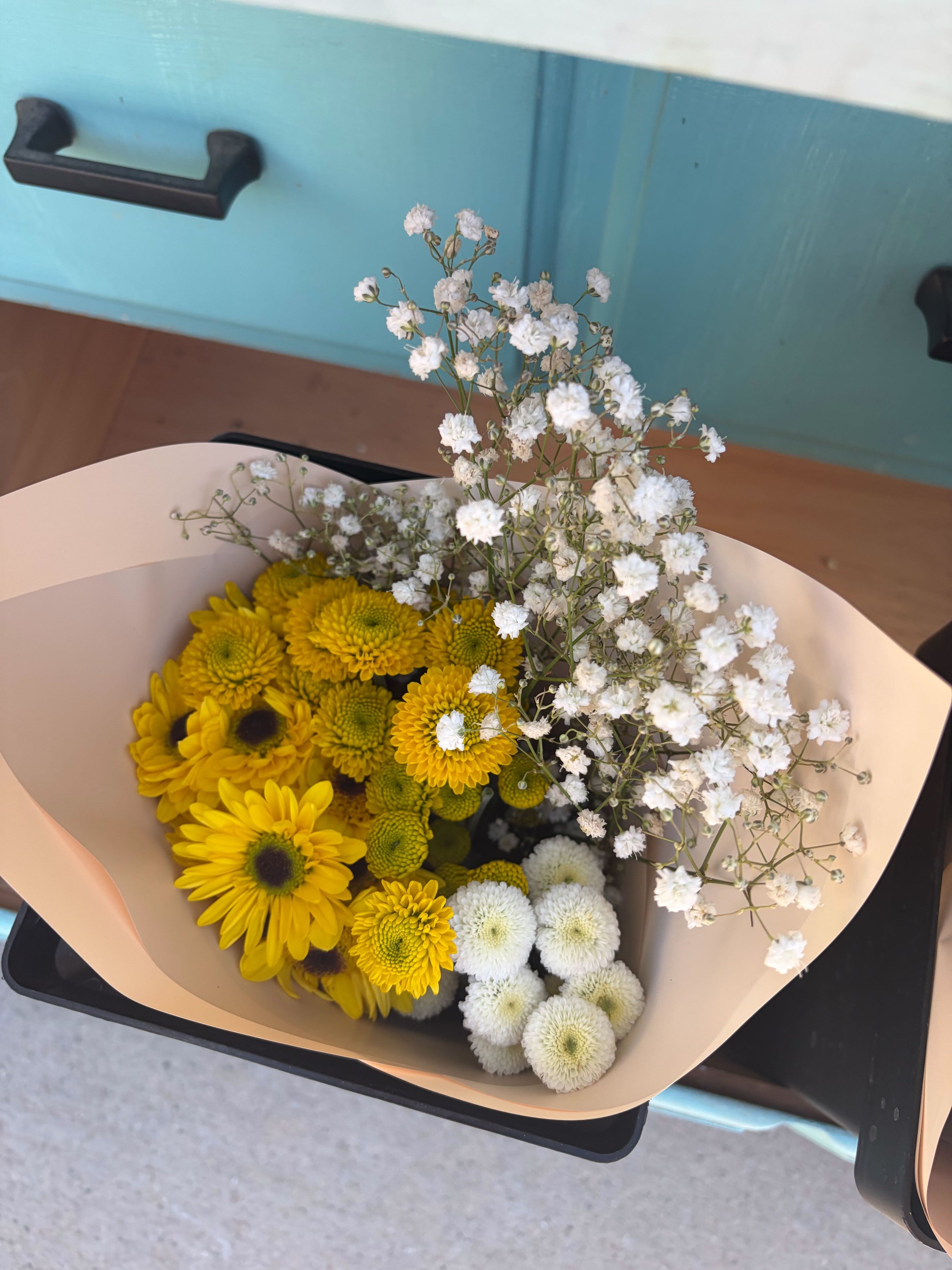 Bouquet of white, yellow, and cream flowers, including daisies and baby's breath, wrapped in cream paper, resting on a black tray.