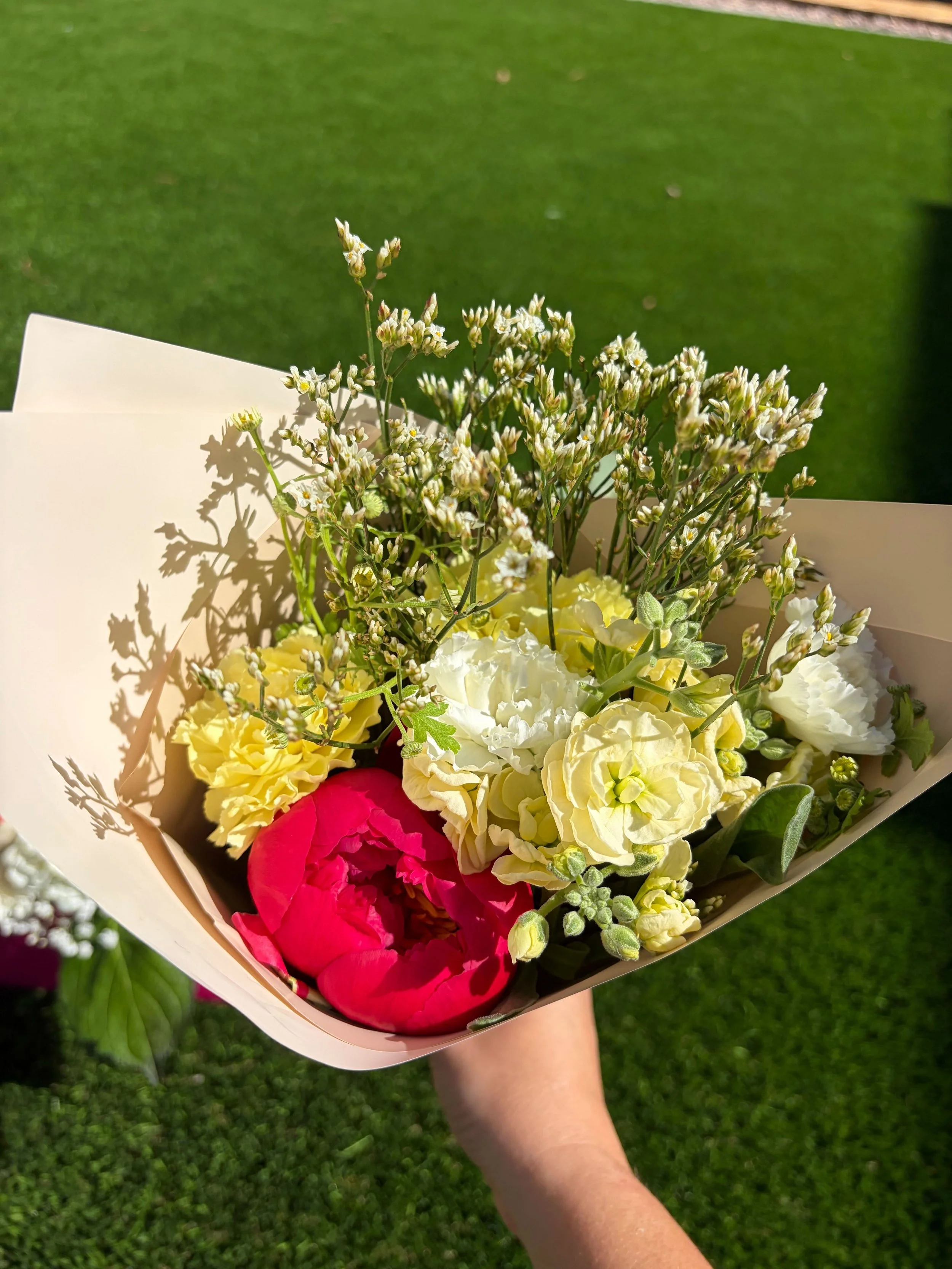 A bouquet of mixed flowers, including white, yellow, and pink blooms, being held by a person outdoors on a grassy background.
