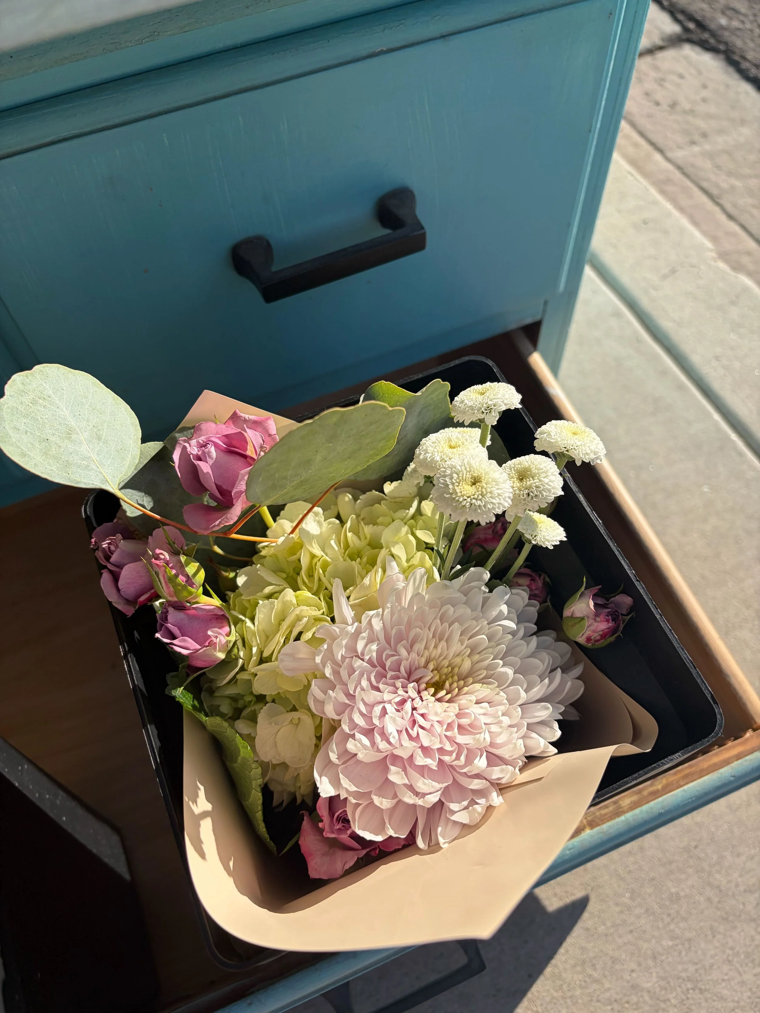 A bouquet of pink, white, and cream-colored flowers in a black container lined with beige paper, placed on a blue metal cart outside near a sidewalk.