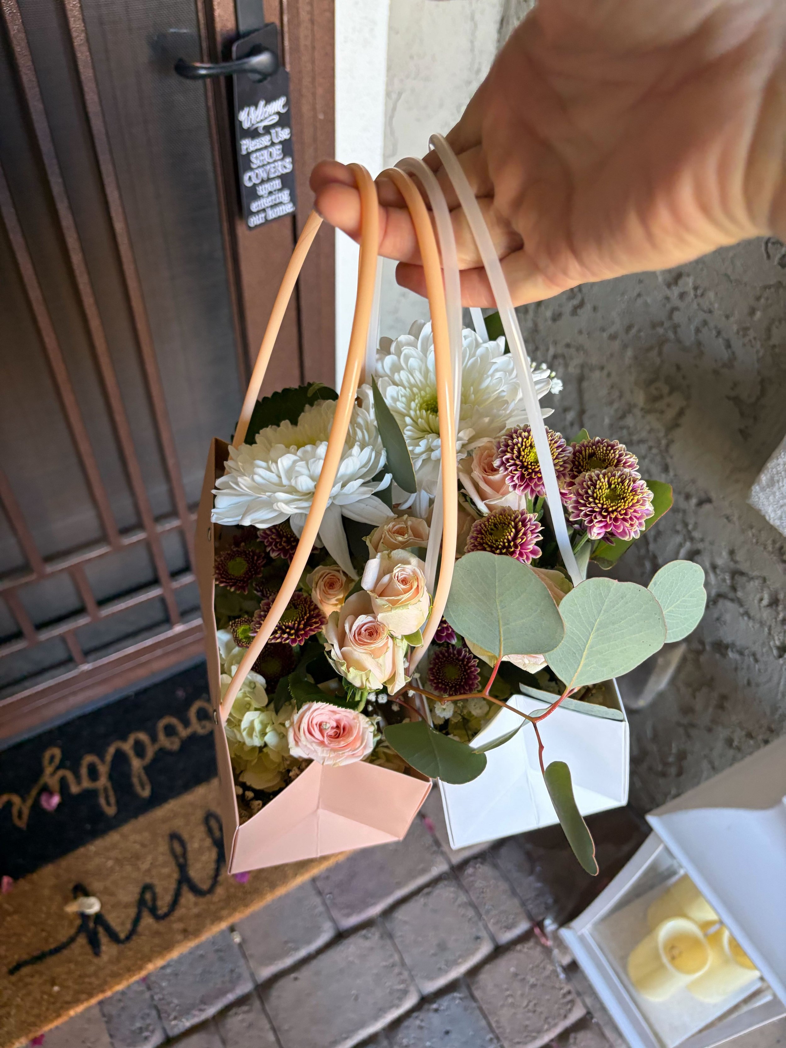 Two flower purses containing white and pink flowers, including chrysanthemums and roses, with green eucalyptus leaves, being delivered to a neighbor's home in Peoria, Arizona.