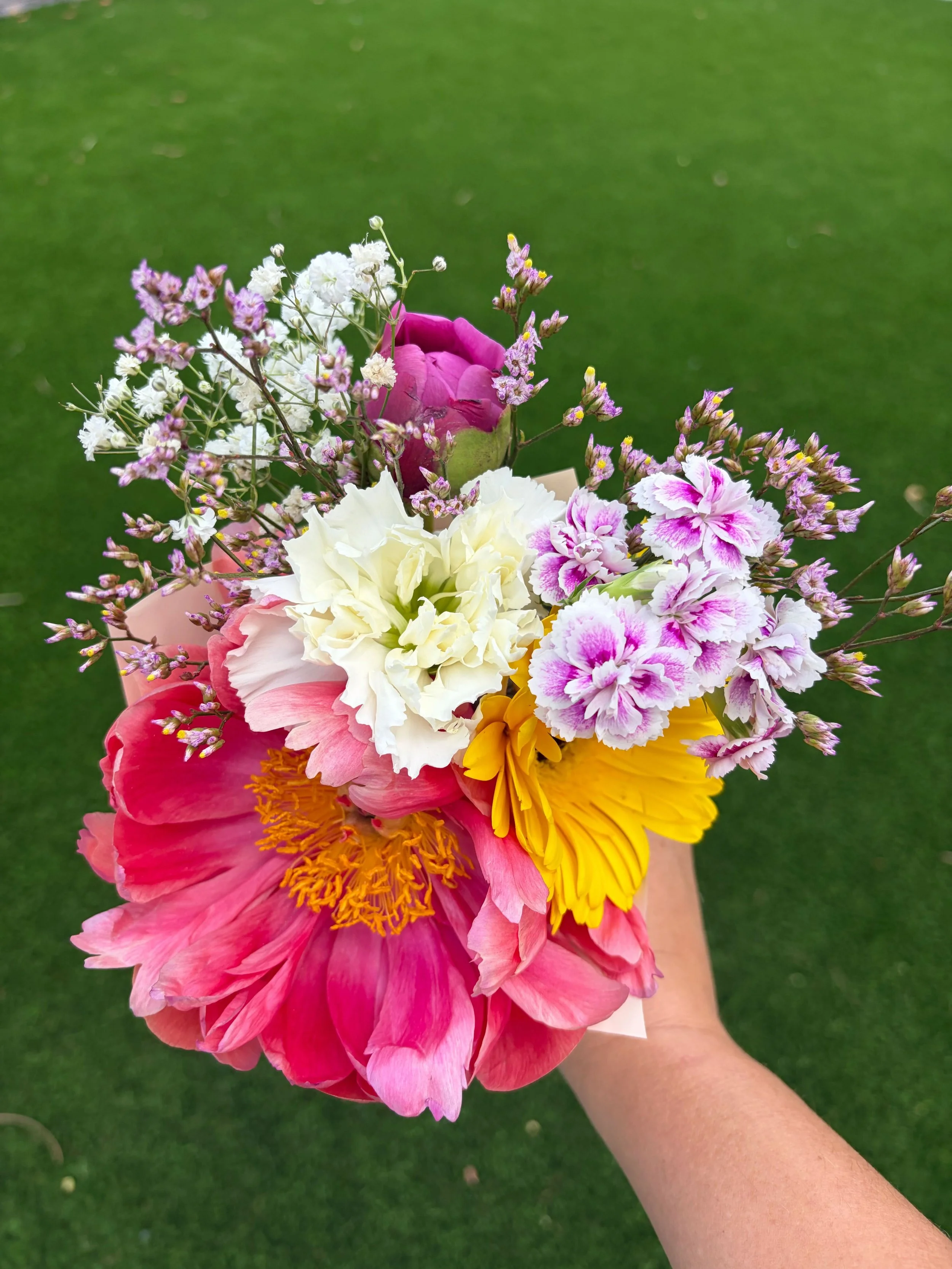 A colorful bouquet of flowers including pink, white, yellow, and purple blooms held by a person's hand against a green grass background.