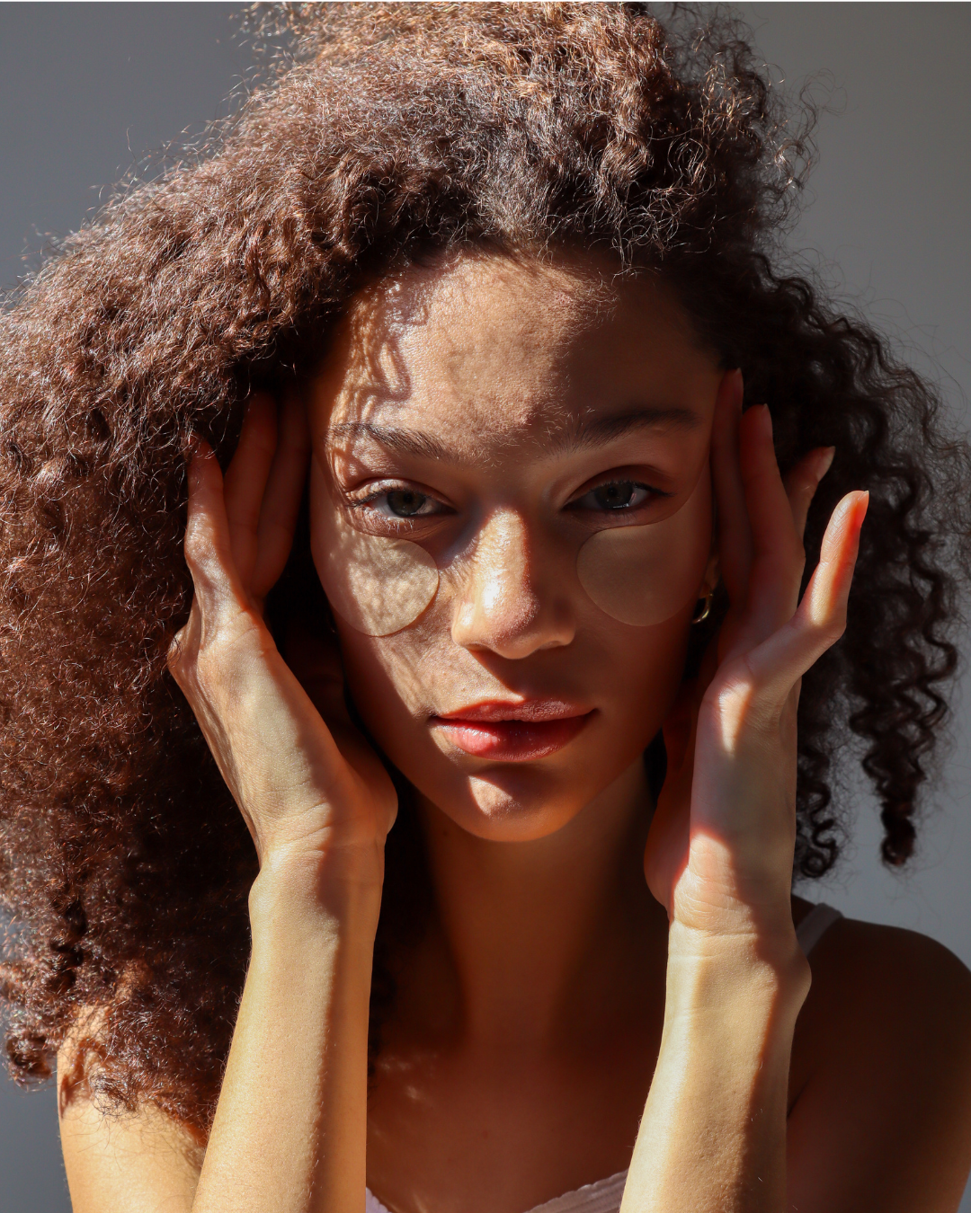 A woman with curly hair holds her head with both hands, sunglasses cast shadow across her face, and sunlight highlights her features.