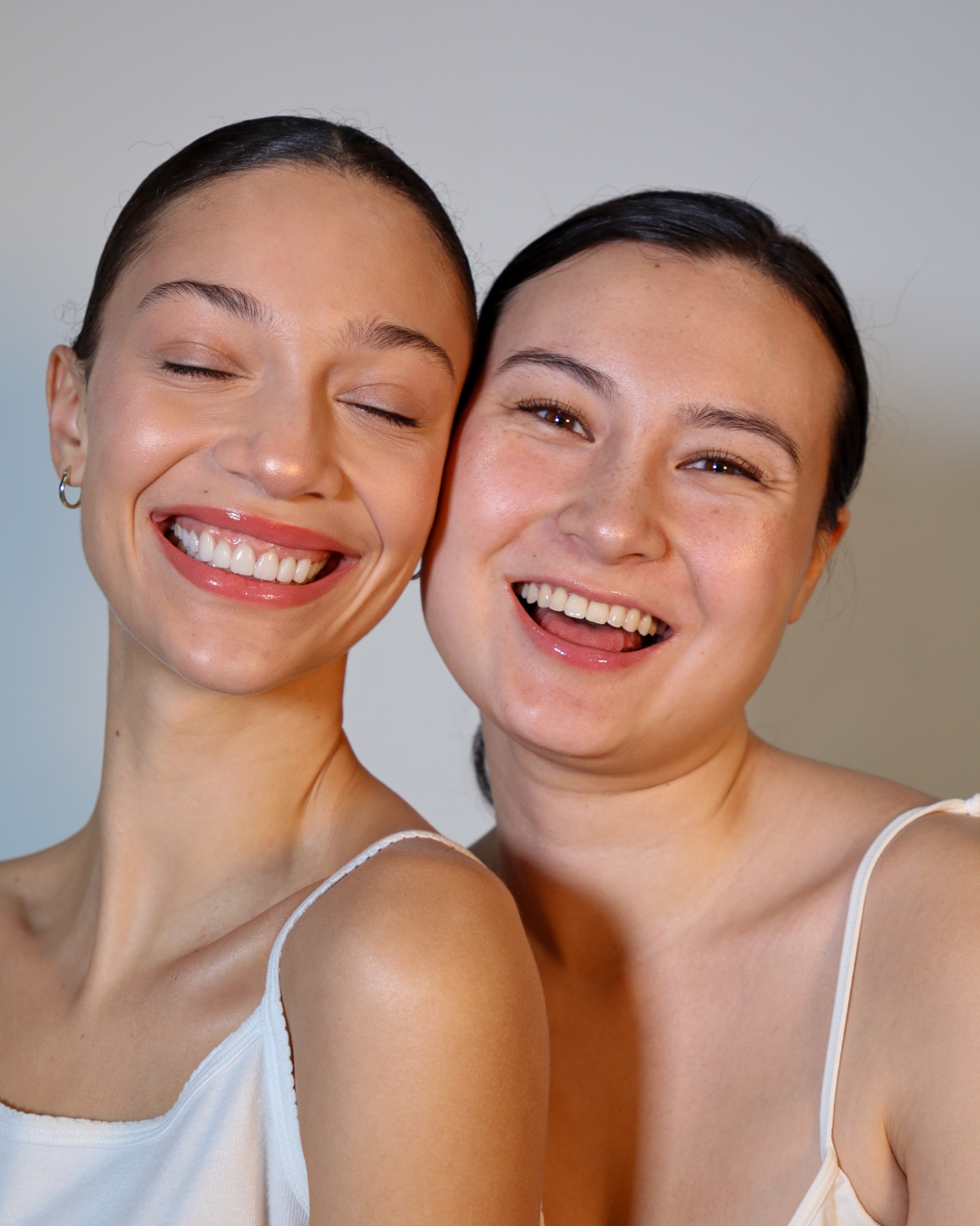 Two women smiling and having fun, one with eyes closed and the other with eyes open, close together in a bright indoor setting.