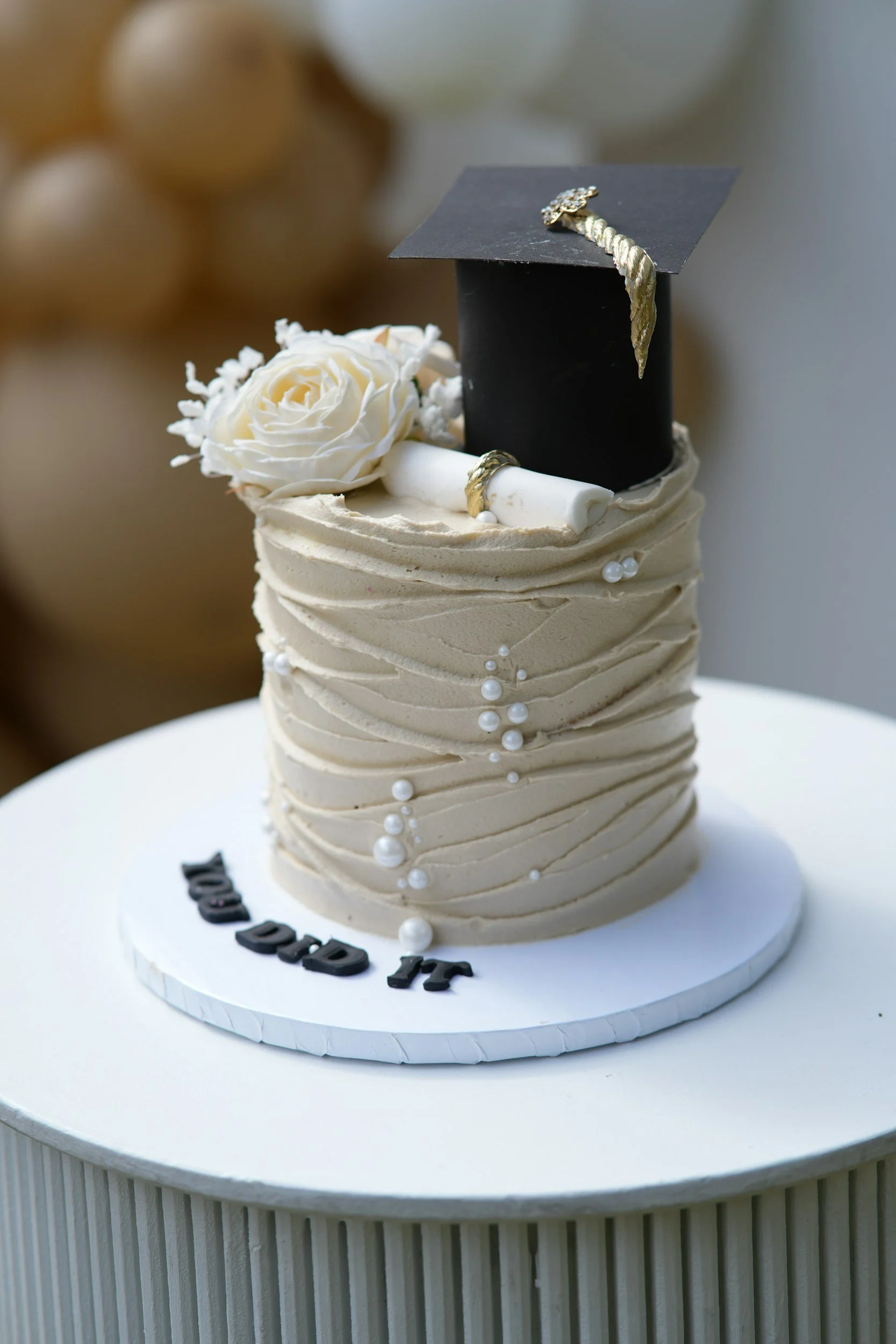 A celebration cake with cream frosting, white rose decoration, and a graduation cap with a diploma and gold embellishments on top. The cake sits on a white round cake board on a table.