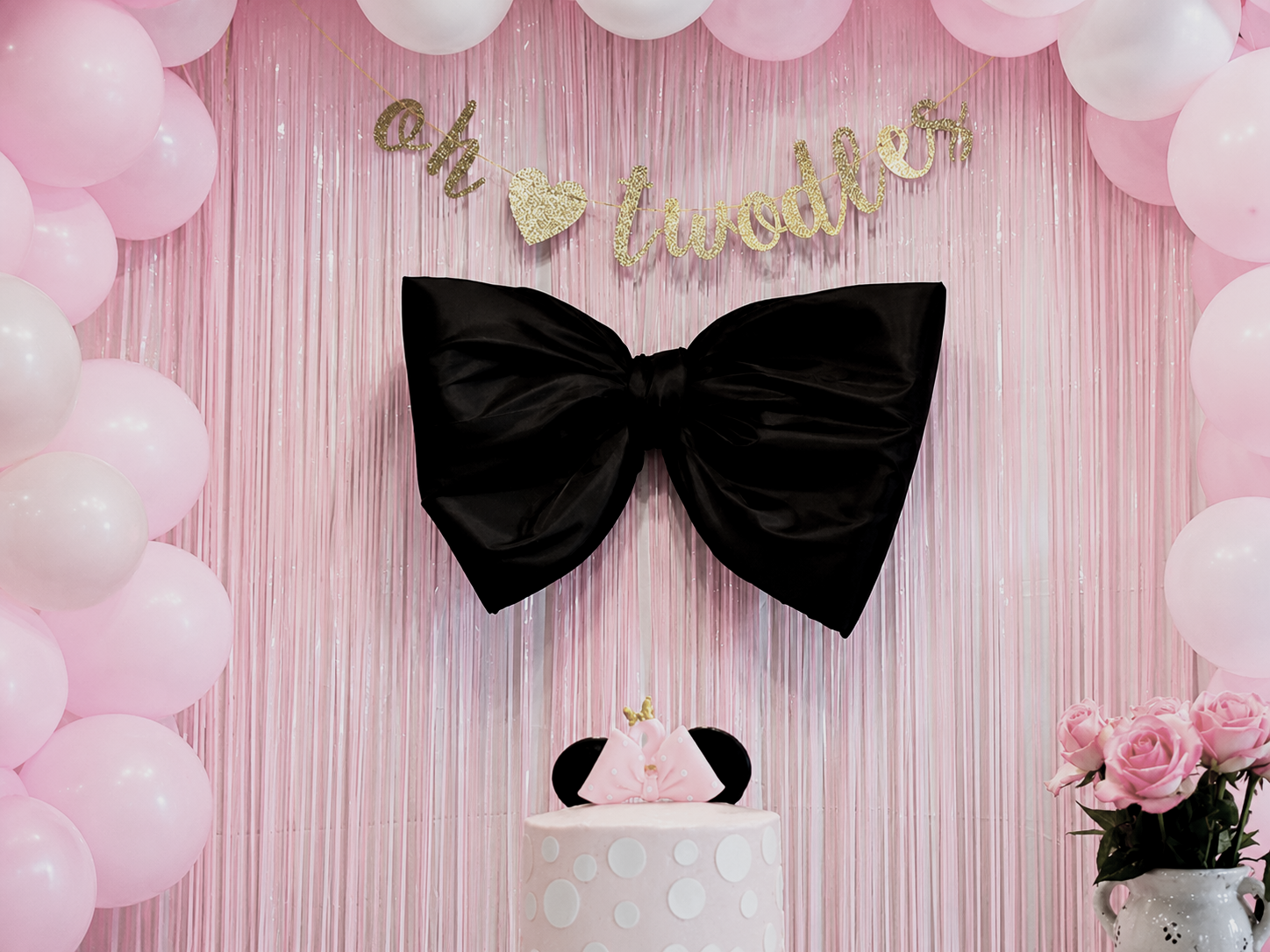 Pink party backdrop with pink balloons, a gold glitter banner saying "oh twoodles," a black bowtie decoration, a pink cake with a Minnie Mouse head silhouette, pink roses, and a pink and white polka dot cake topper.