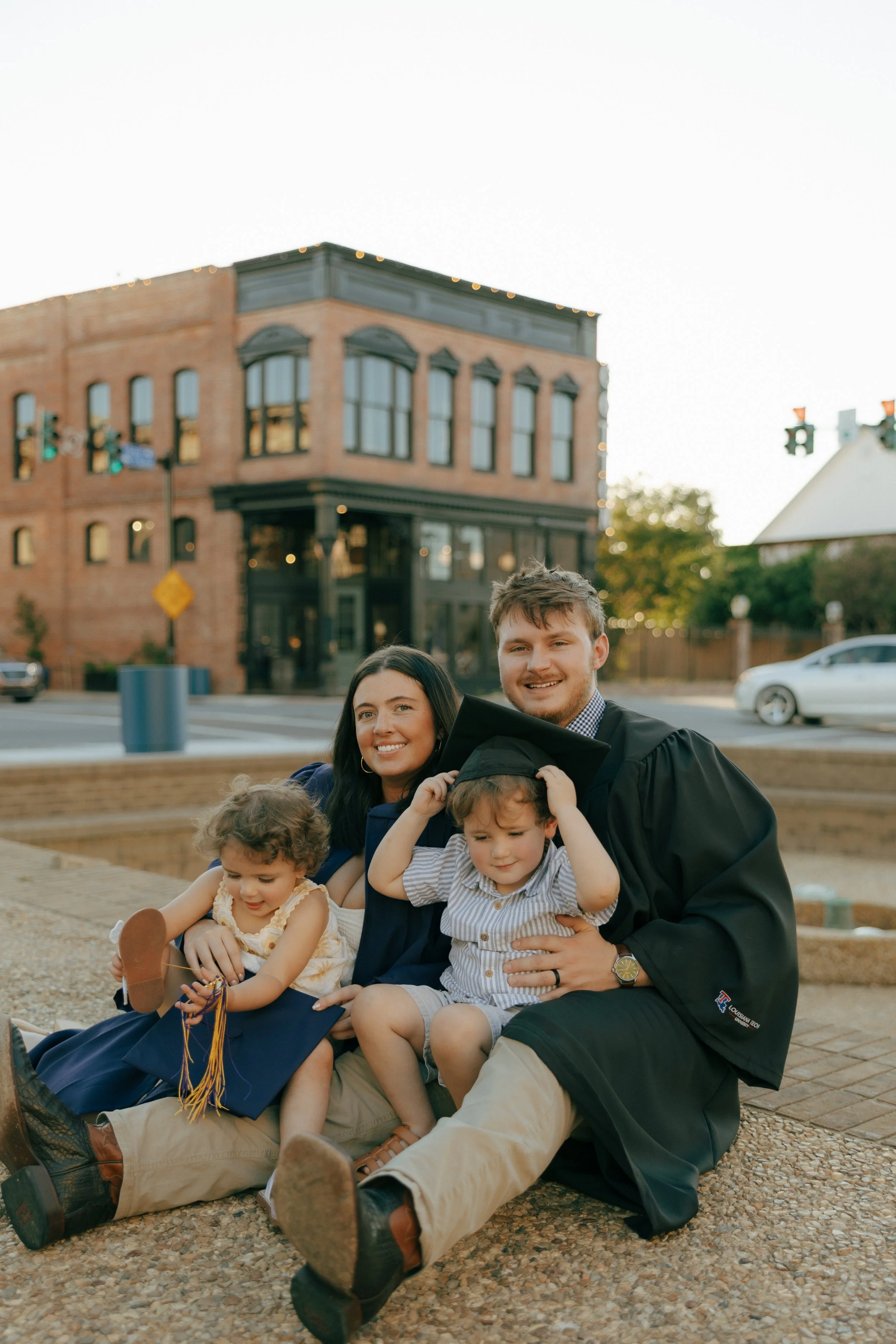 A group of family members celebrating a graduation outdoors, with a brick building and traffic lights in the background.
