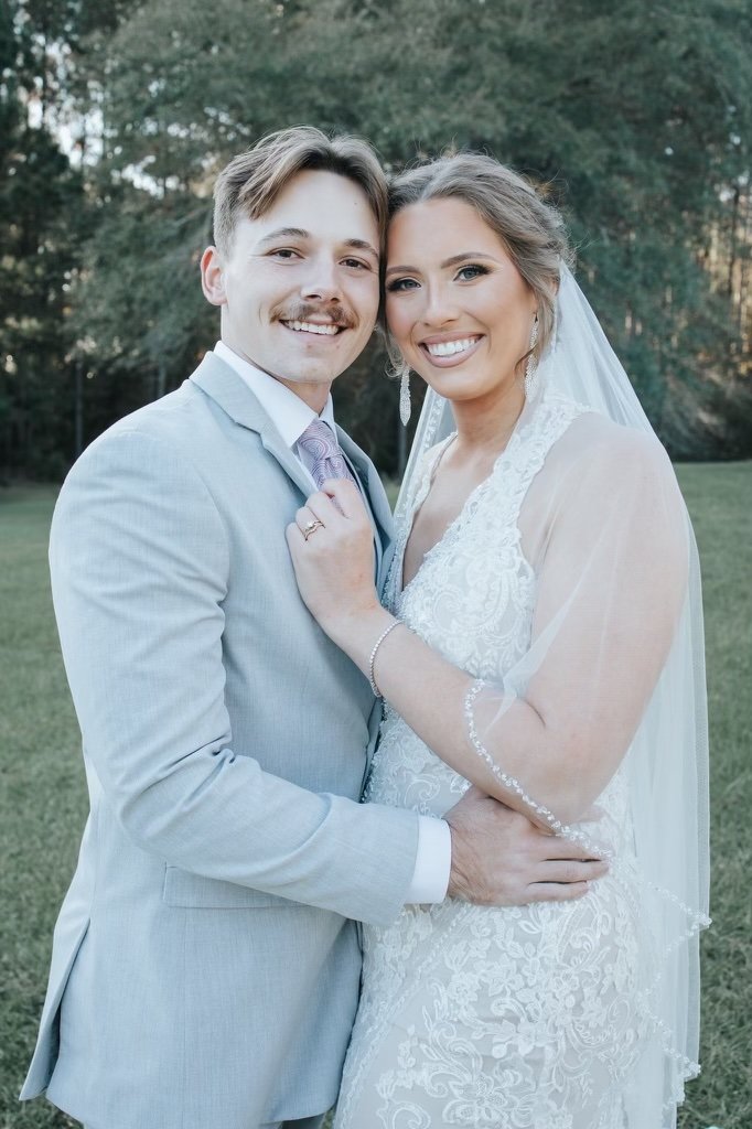 A newlywed couple smiling outdoors, the groom in a light gray suit with a lavender tie, and the bride in a lace wedding gown with a veil, embracing each other with trees in the background.