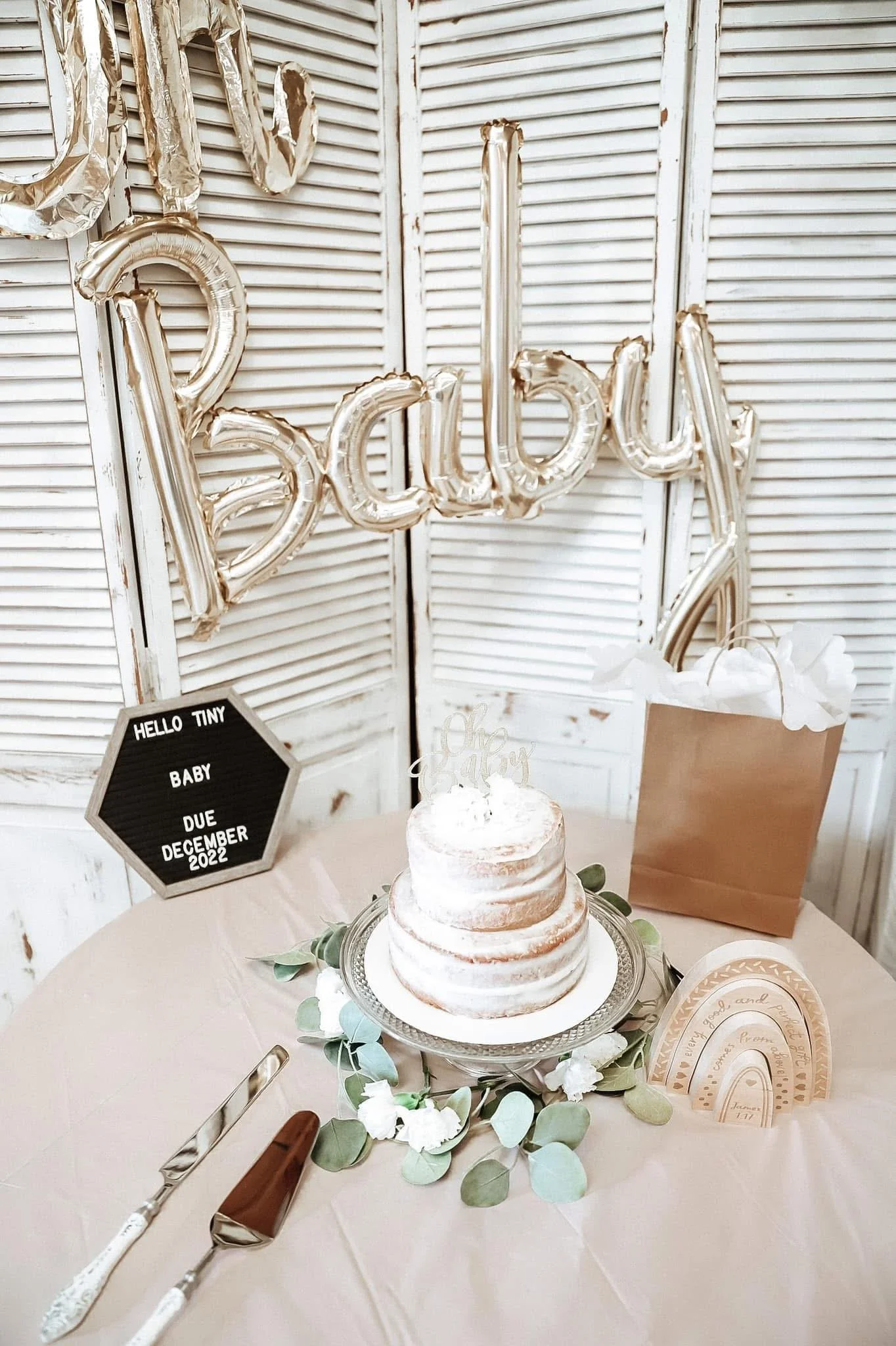 Decorative celebration setup featuring a tiered naked cake with white frosting and green leaves, gold and silver balloons spelling 'Baby', a black sign with white letters indicating a due date, white flowers, and a gift bag, all against a white paneled background.