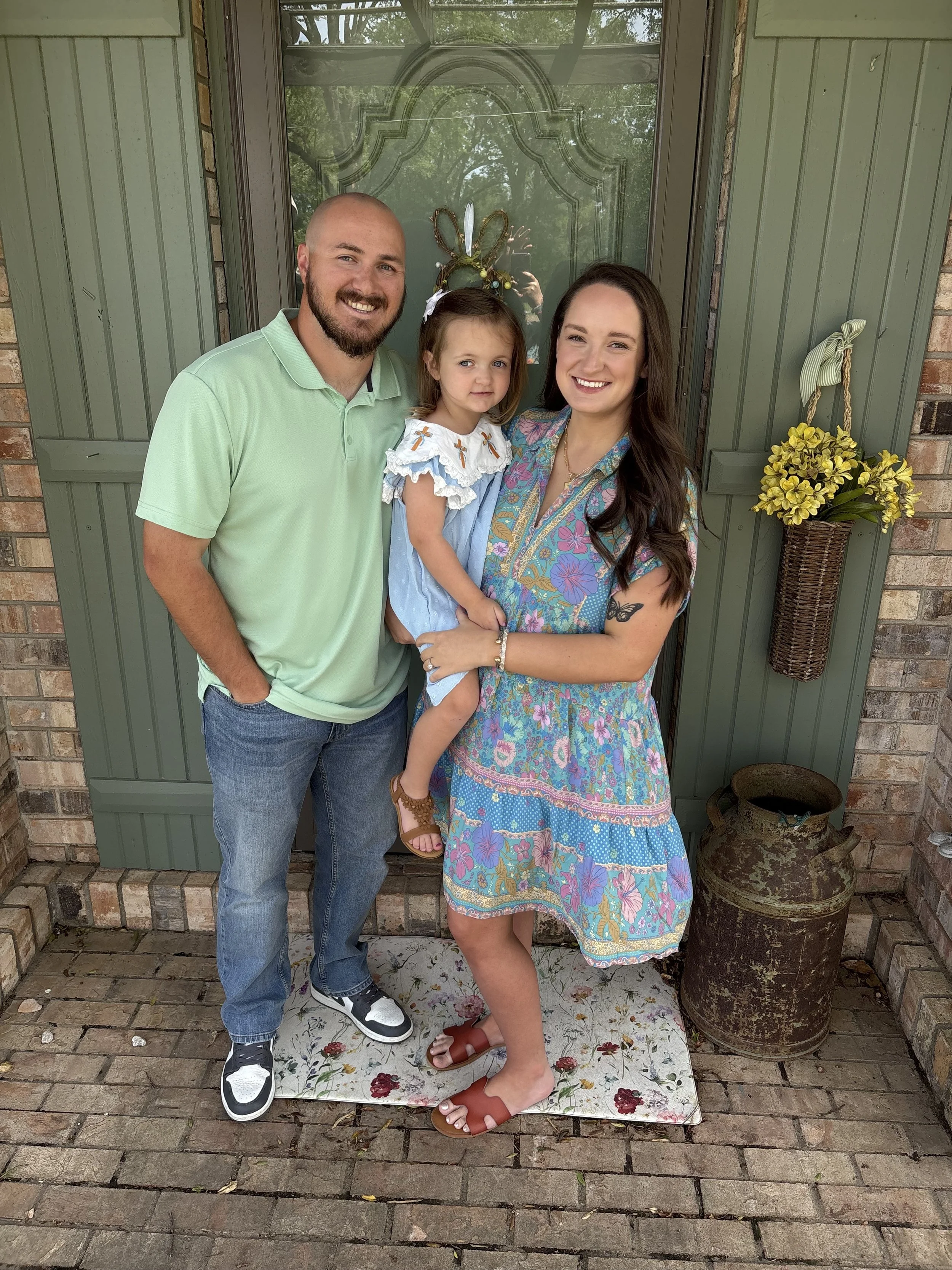 A smiling family of three, including a man, woman, and young girl, stands on a brick porch in front of a green door with a glass window featuring decorative designs. The girl wears a headband with bunny ears and a dress with crosses, while the woman wears a colorful floral dress and brown sandals, and the man wears a light green polo shirt and jeans. The porch has a floral rug, a flower basket on the wall, and an antique water jug on the ground.