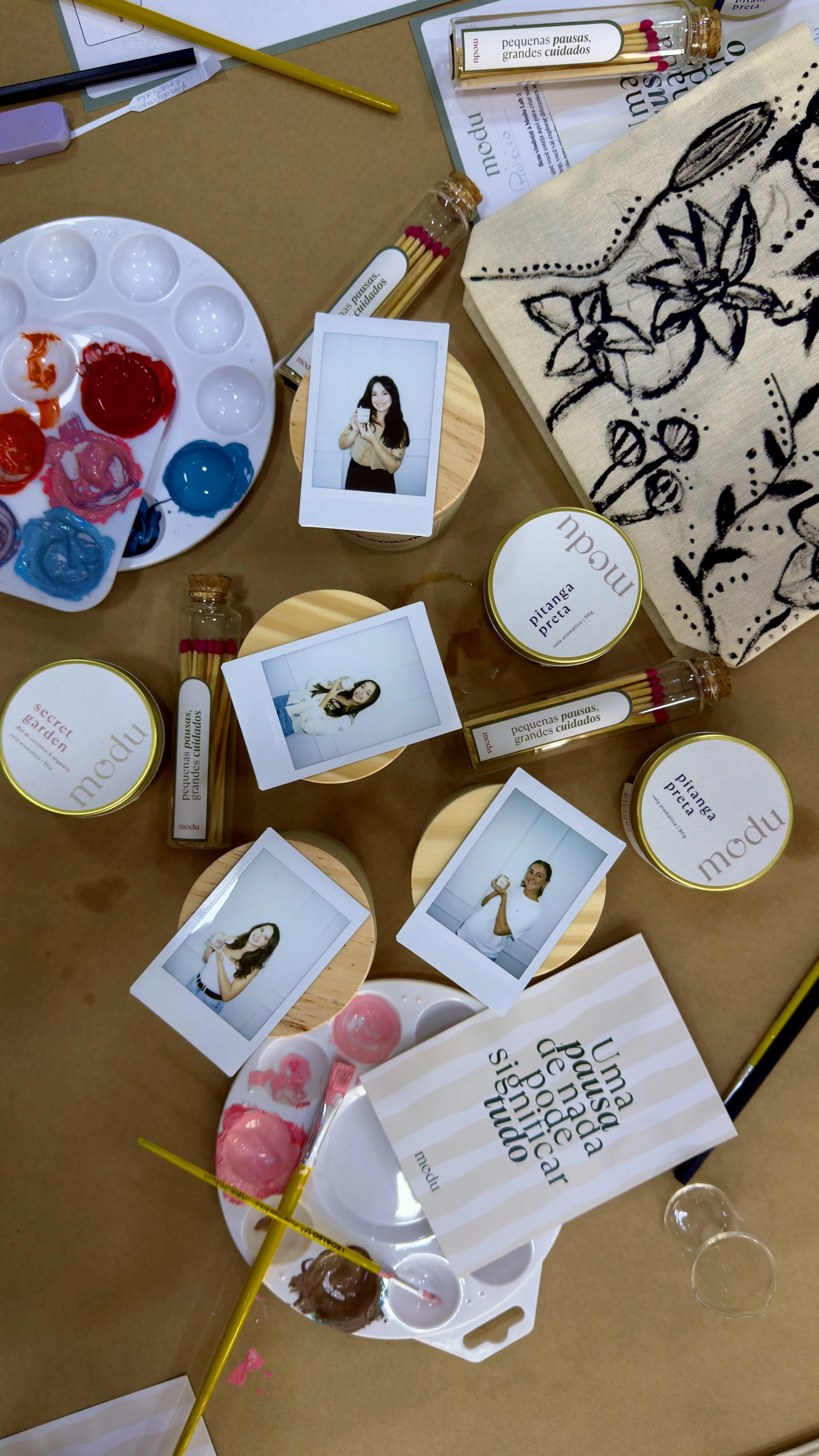 Tabela de pintura com tintas coloridas, pincéis, caixas de potes com味 de tinta e fotos de pessoas, além de um tecido com estampa de folhas e flores