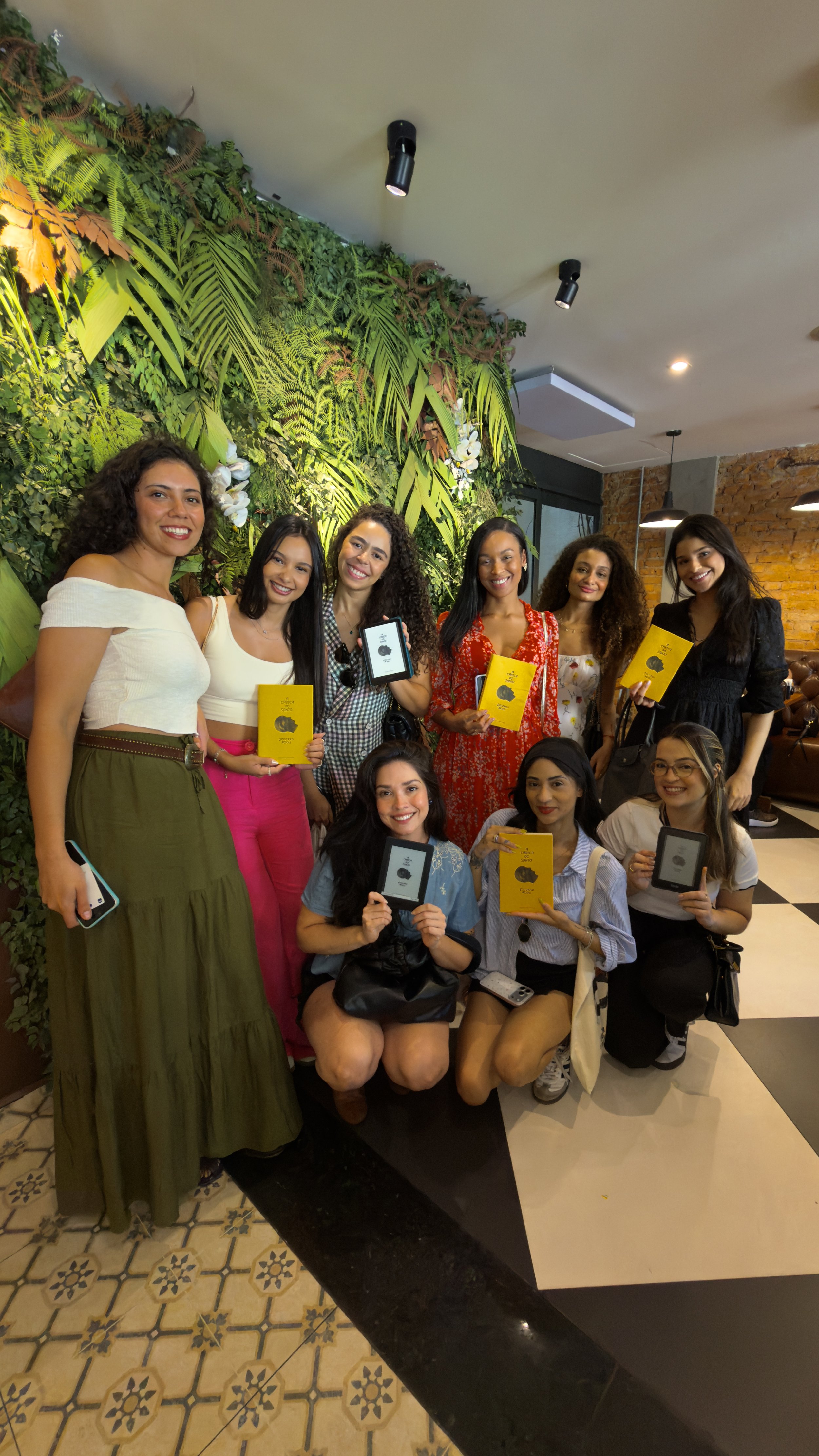 Grupo de mulheres sorrindo, algumas segurando livros ou e-readers, em ambiente decorado com plantas e iluminação moderna.