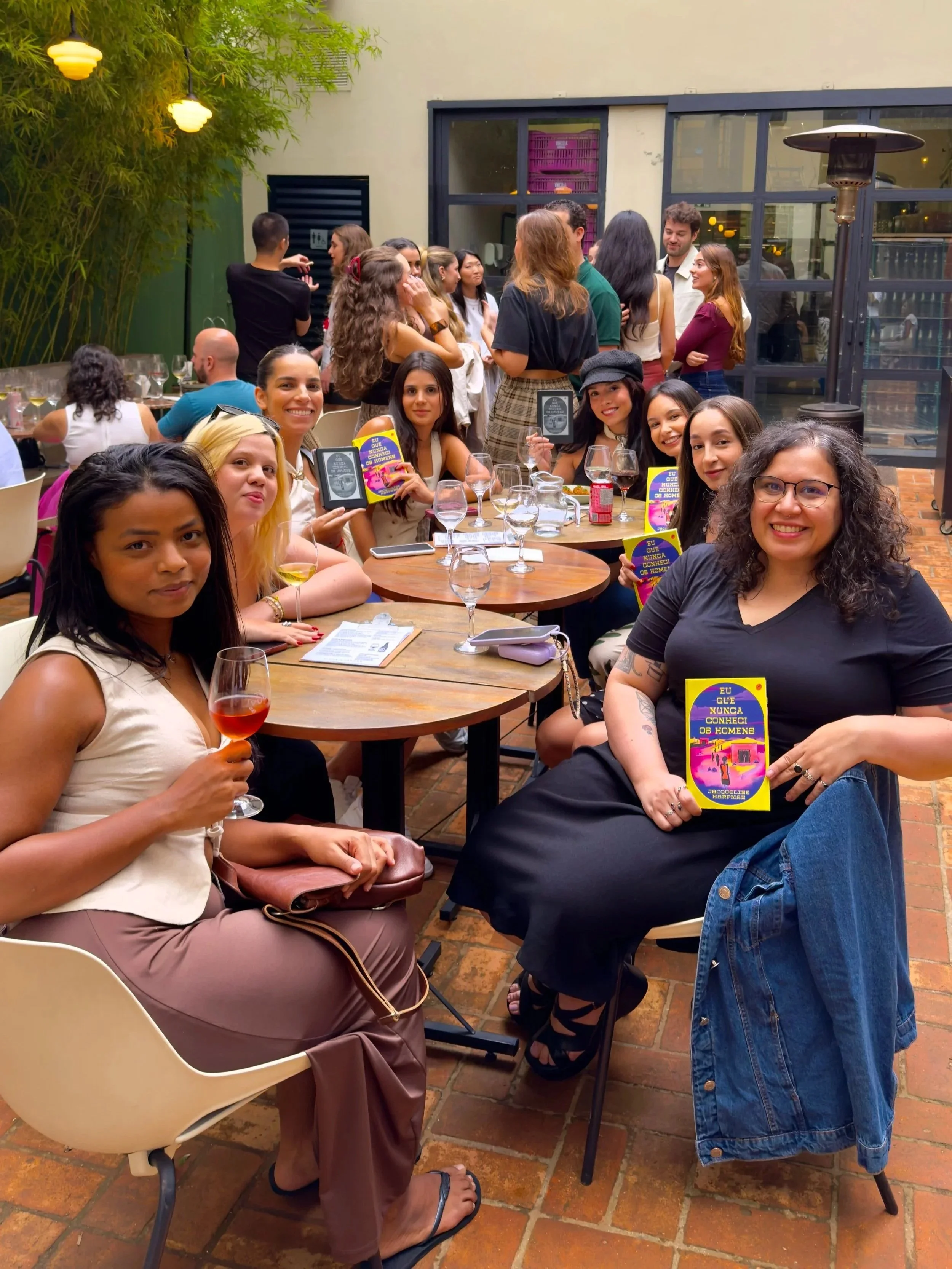 participantes do sentimental clube durante o clube do livro, ao redor de uma mesa em um restaurante, algumas segurando copos e livros, outros conversando, com um ambiente bem iluminado, plantas verdes decorativas e janelas grandes ao fundo.