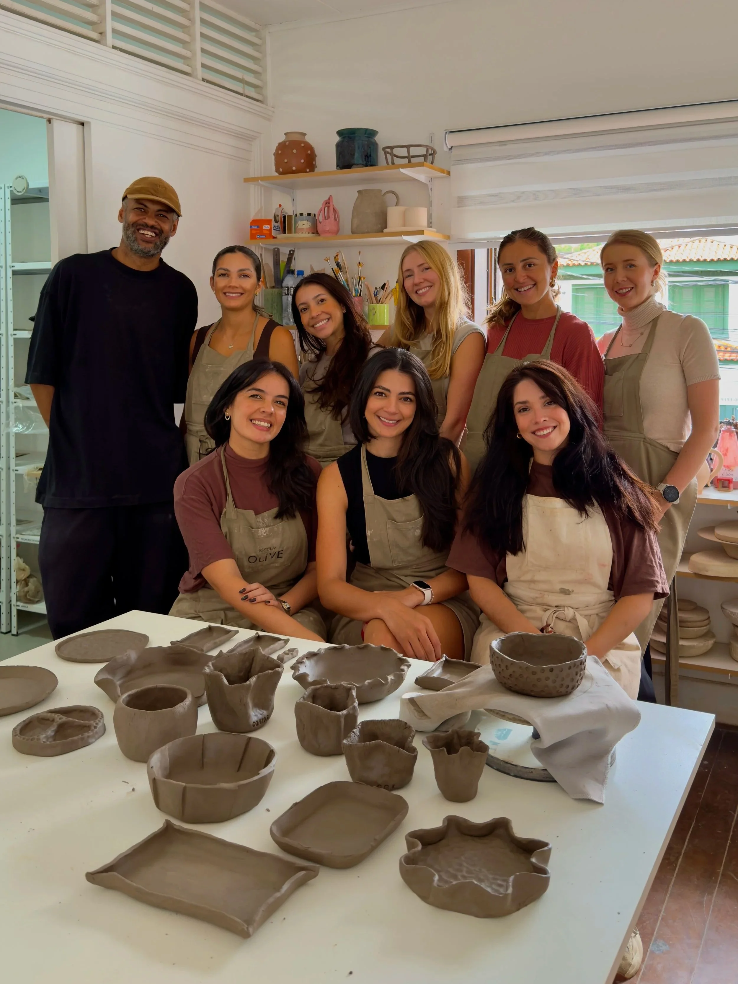 Grupo de pessoas sorrindo em volta de uma mesa com várias peças de cerâmica em processo de confecção em uma oficina de cerâmica.