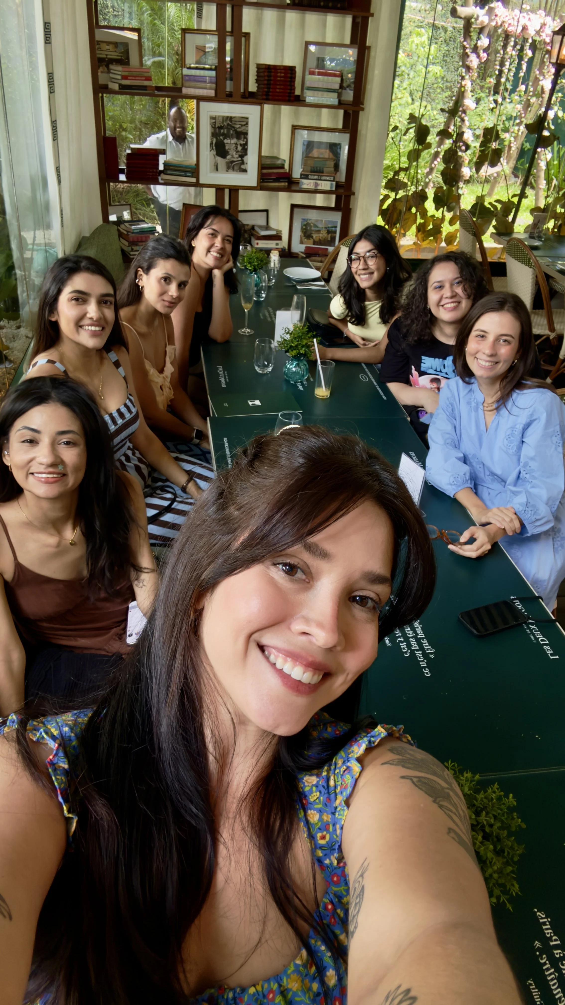 Grupo de mulheres sorrindo, sentado em uma mesa de restaurante ou café, com uma parede de janelas ao fundo cheia de plantas e um homem visível na porta ao fundo.