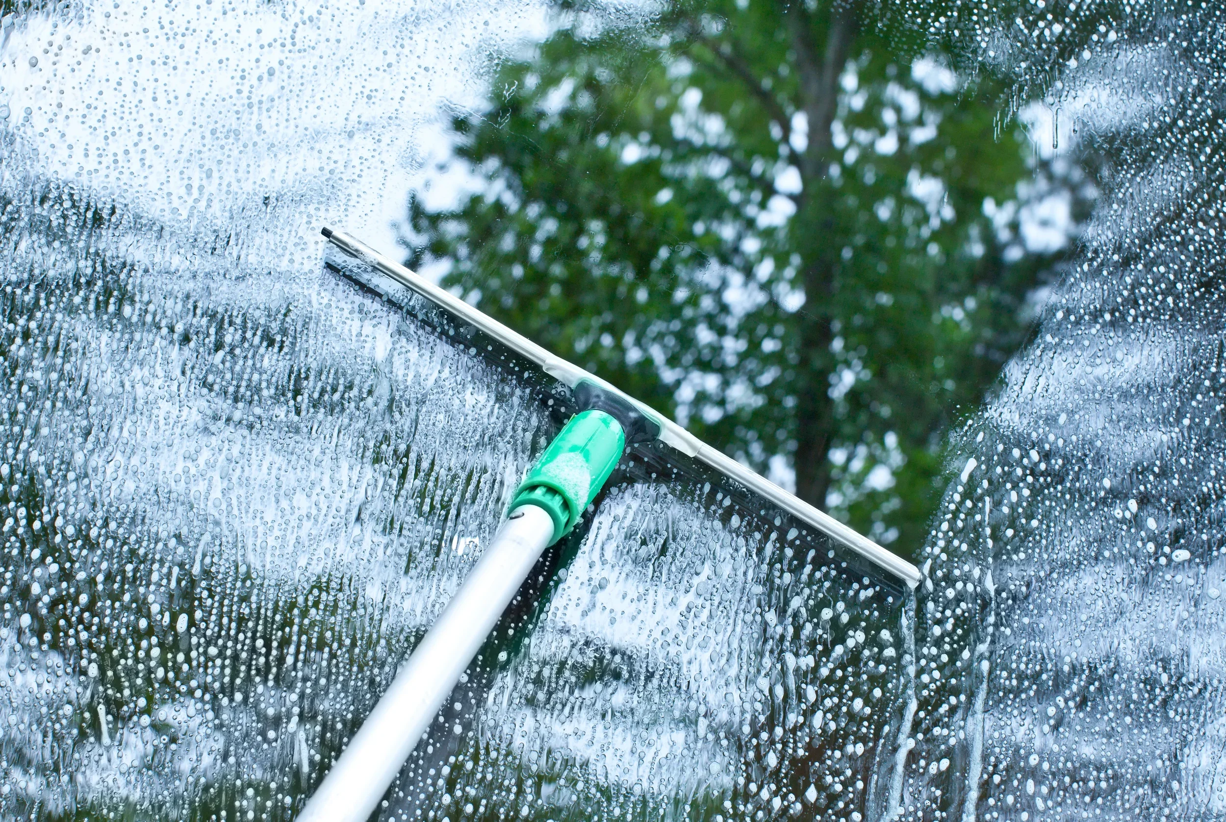 A windshield being cleaned with a squeegee on a rainy day, with water droplets on the glass and greenery visible through the window.