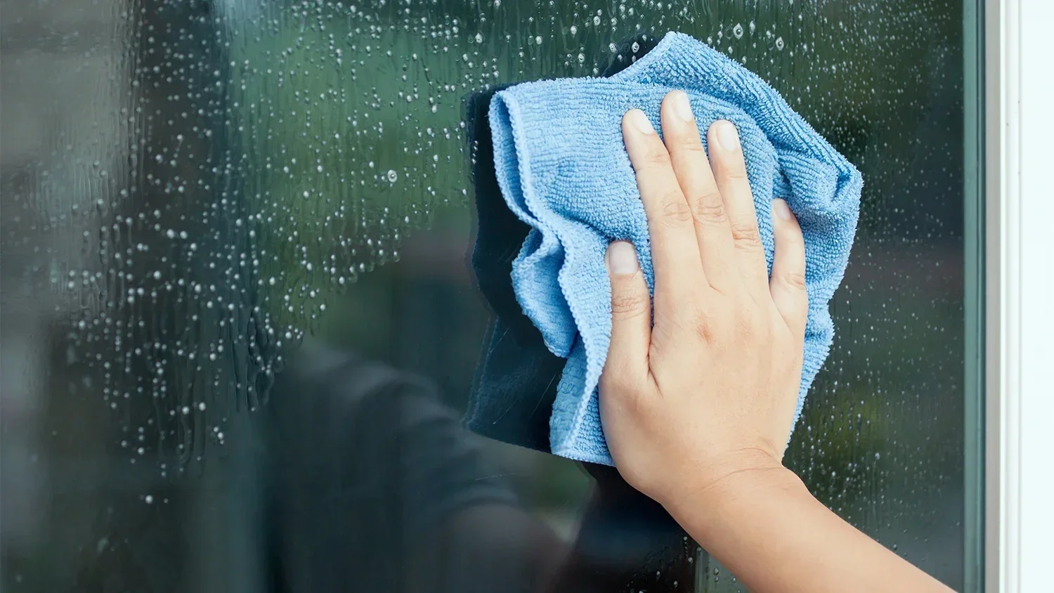 A person cleaning a glass window with a blue microfiber cloth, removing soap or dirt, with water droplets on the glass.