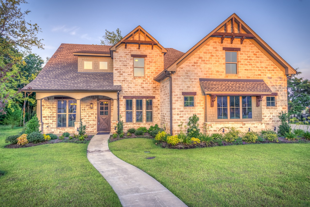 A two-story house with stone and stucco exterior, front yard with green grass, landscaping, and a curved sidewalk leading to the front door, under a clear blue sky.