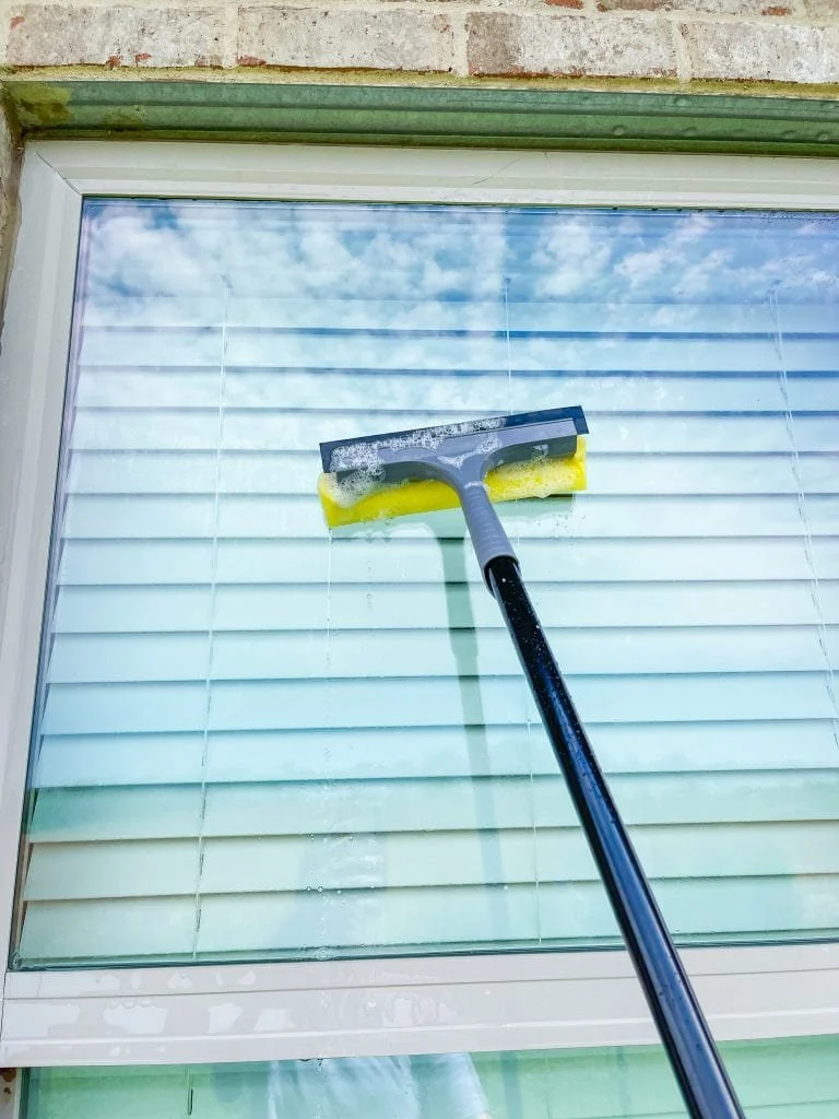 A window with closed blinds being cleaned with a squeegee and yellow scrubber, seen from inside a building.