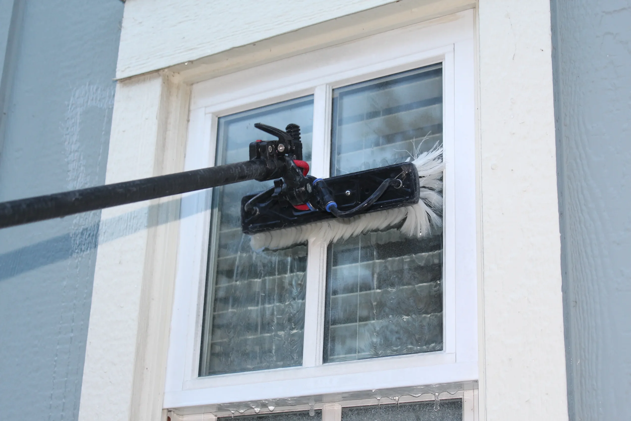 A window cleaning tool attached to a window, with the tool having a large white scrubber and black handle, cleaning the window on a white-framed window with curtains inside.